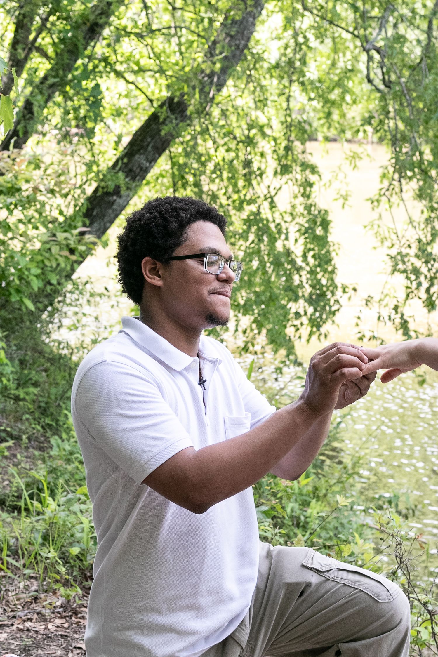 Young man with glasses in white polo shirt smiling outdoors near a pond, reaching out to receive something.