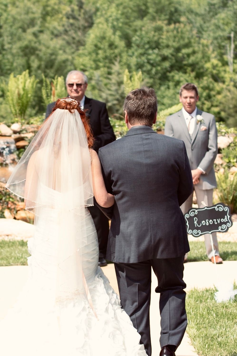 Bride walking down the aisle with escort at outdoor wedding ceremony, groom waiting at altar.