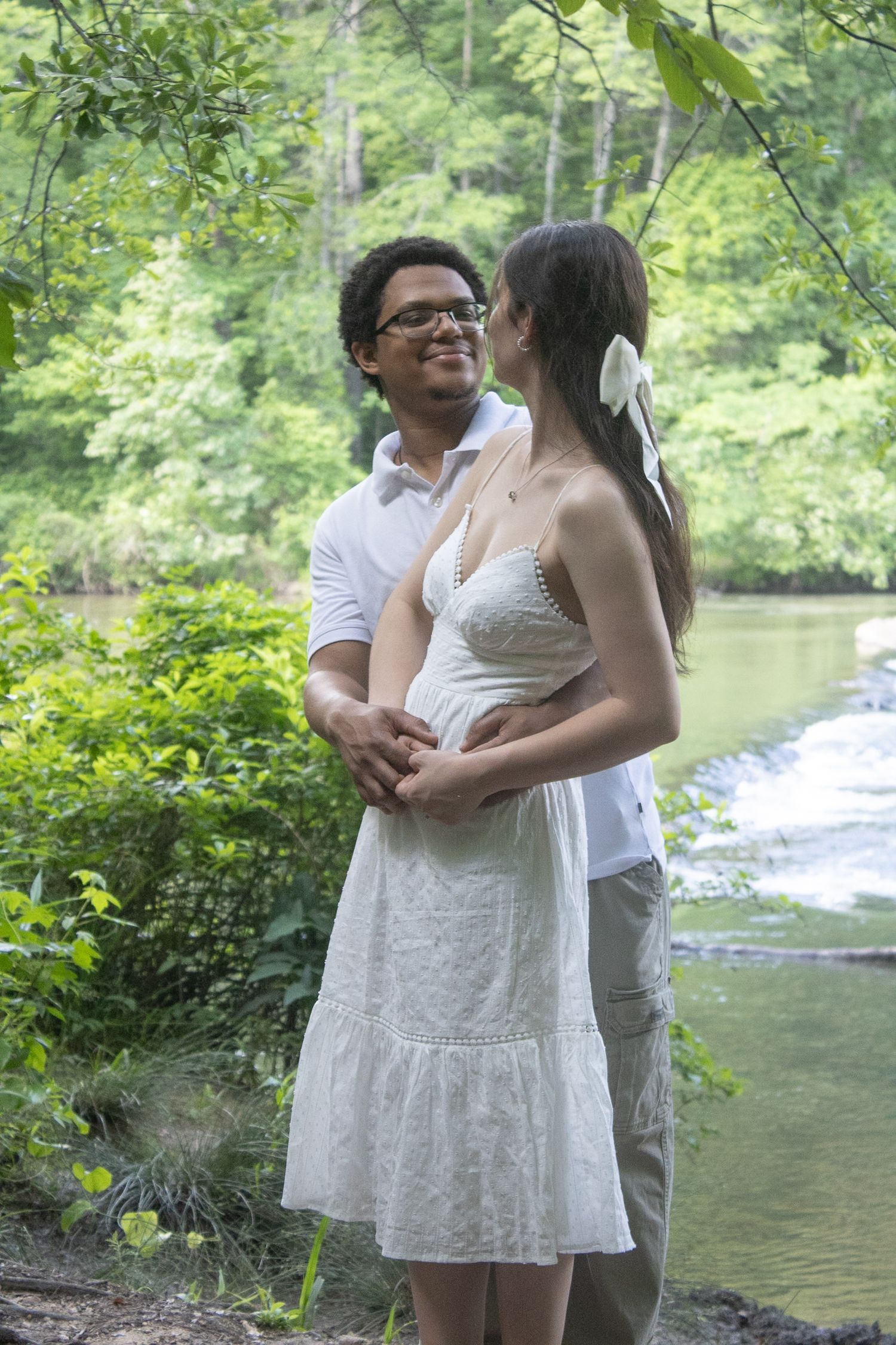 Couple in love embracing outdoors near a serene waterfall, woman in white dress, lush green trees in background.