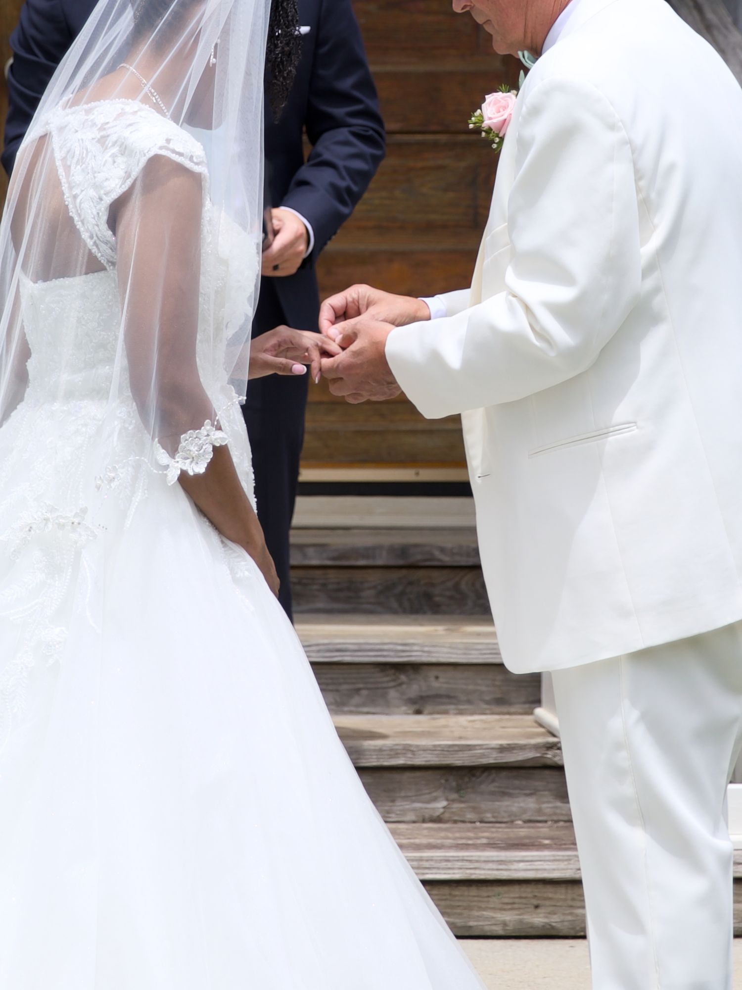 Groom in white suit placing wedding ring on bride's finger during outdoor ceremony.