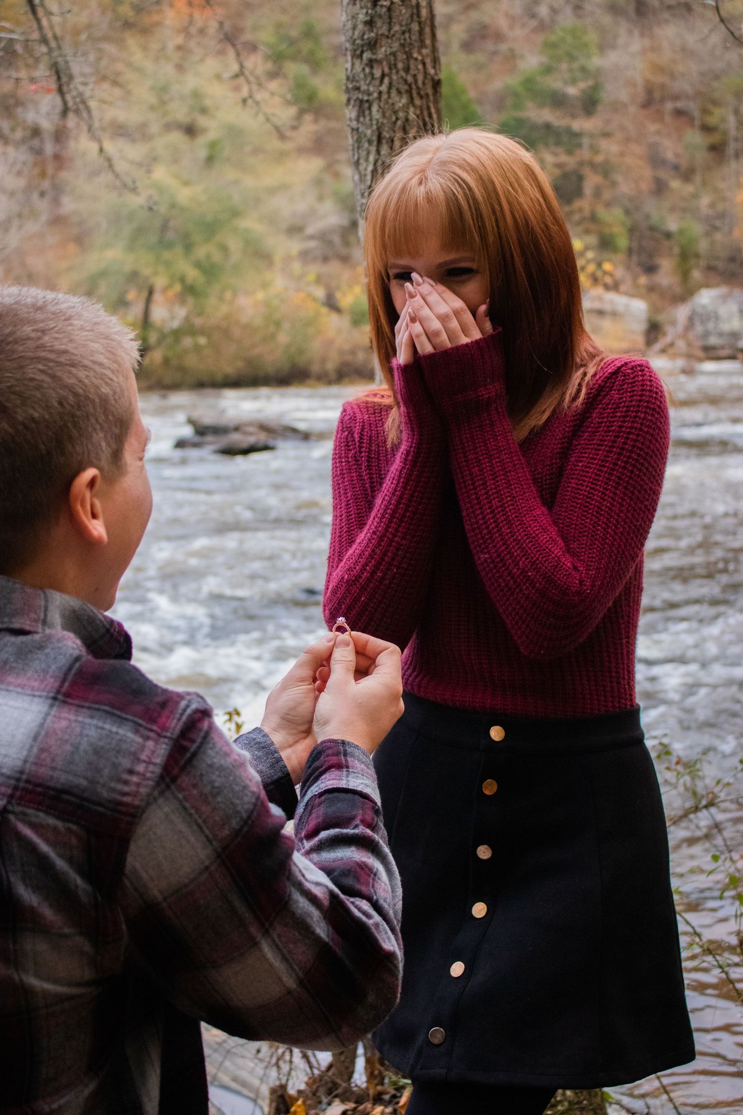 Man kneeling to propose to emotional redhead woman in pink sweater by a river in autumn forest.
