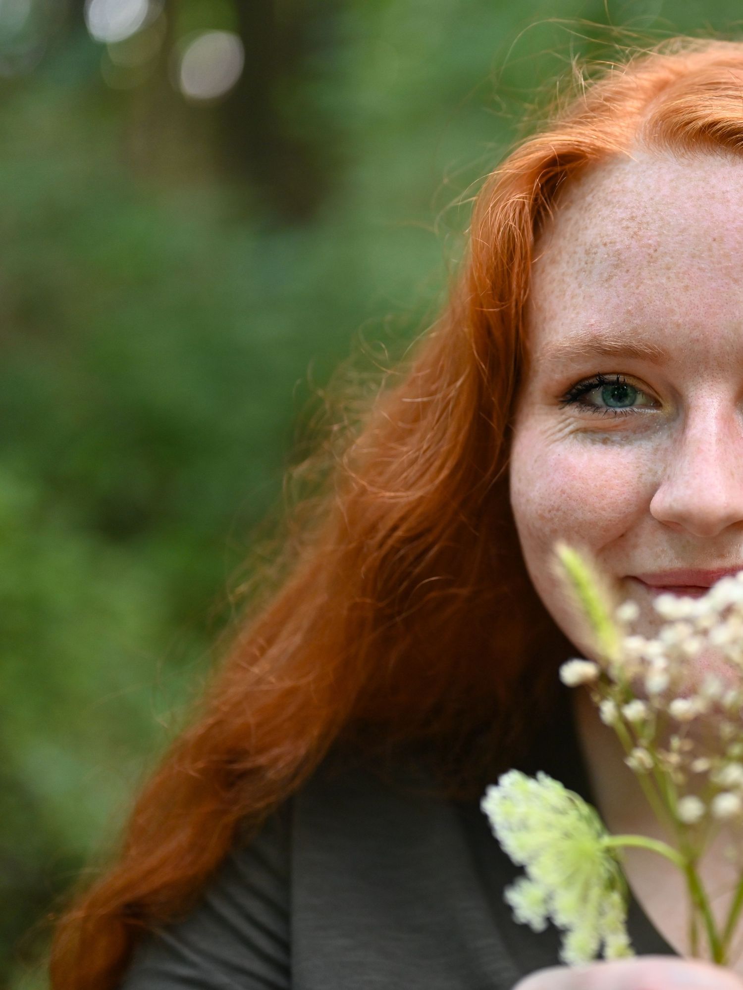 Young woman with long red hair and freckles smiling while holding white wildflowers outdoors.