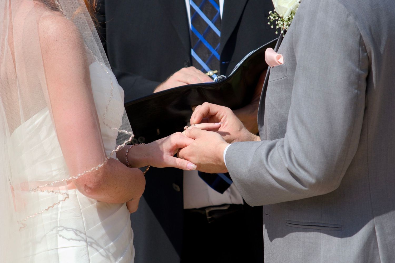 Officiant placing wedding ring on bride's finger during outdoor ceremony, groom in gray suit stands nearby.