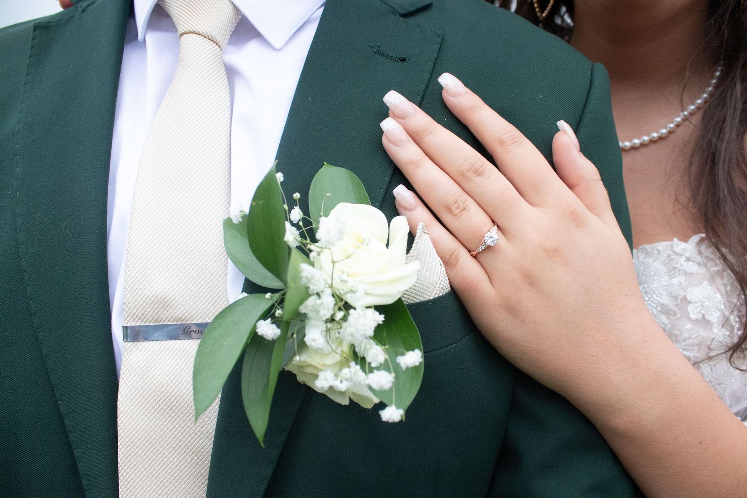 Close-up of bride showing diamond ring resting hand on grooms green suit boutonniere.