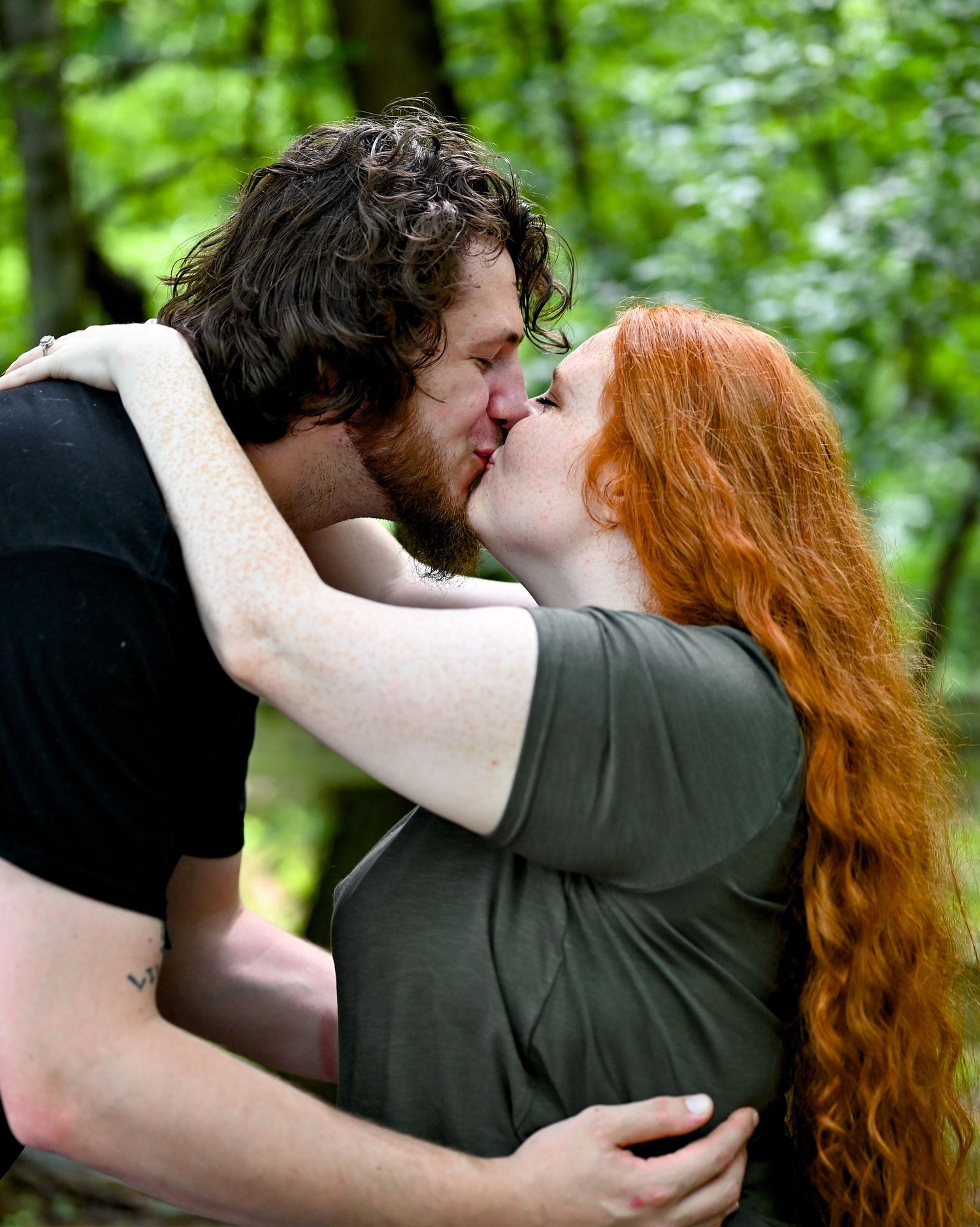 Couple sharing a romantic kiss outdoors in a lush green forest, man with dark curly hair and woman with long red hair.
