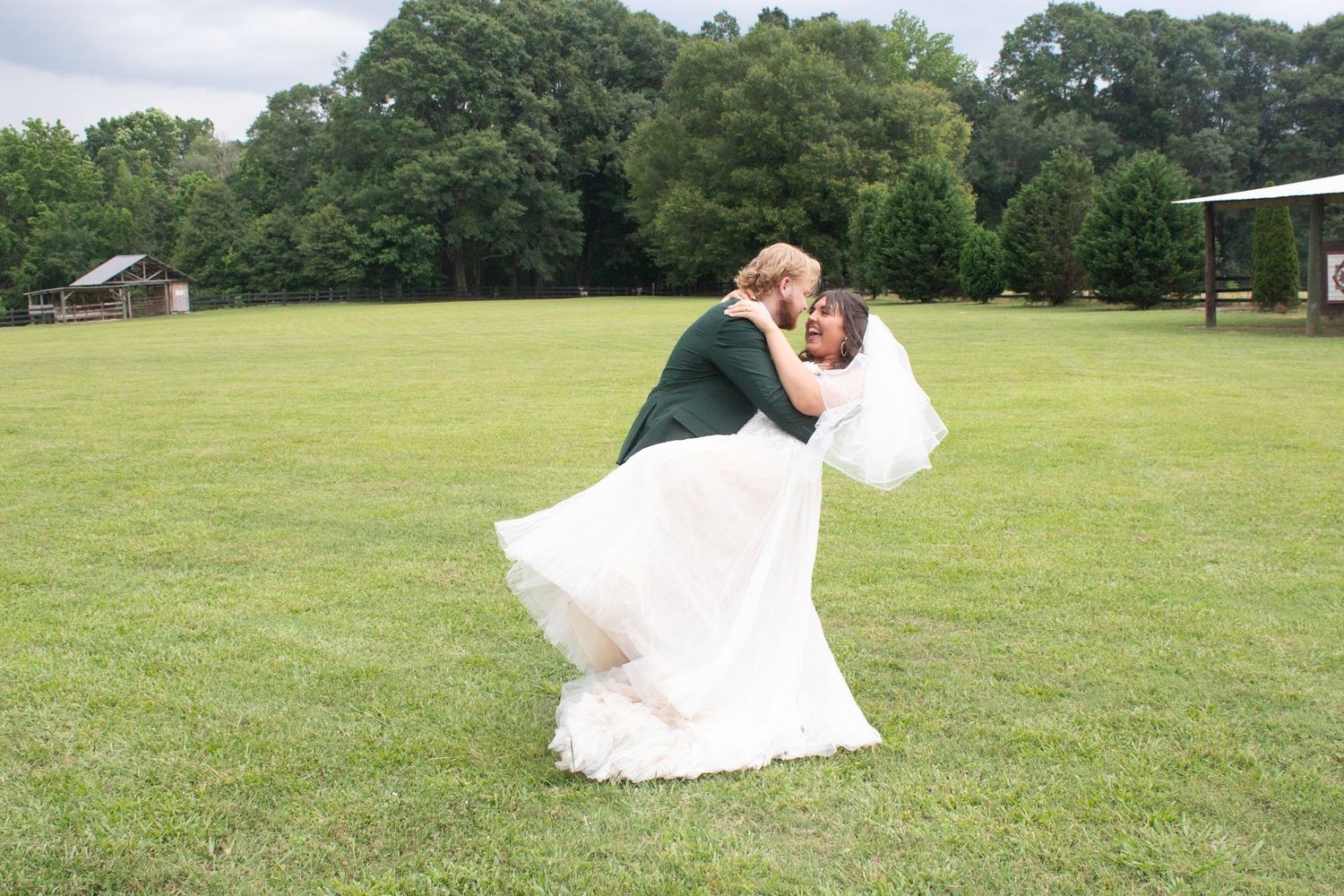 Groom dipping bride in white gown for romantic kiss on lush green lawn at outdoor wedding ceremony.