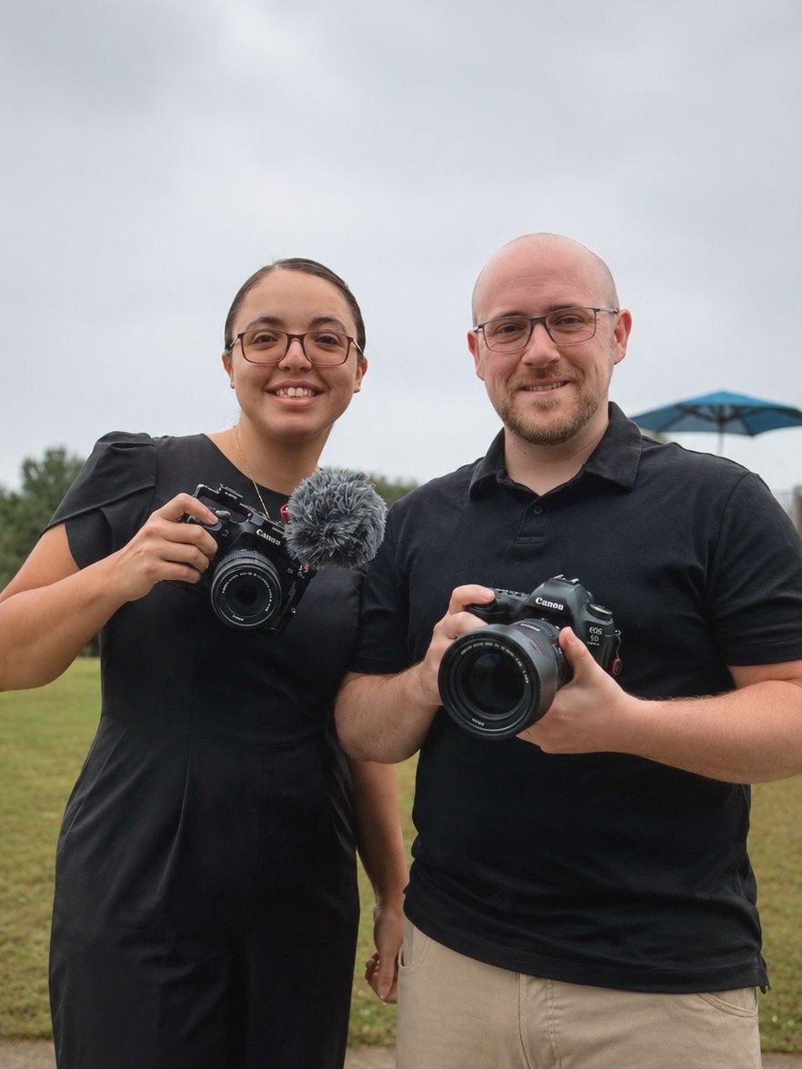 Two photographers smiling outdoors holding DSLR cameras, woman in black dress with microphone attachment, man in black polo shirt.