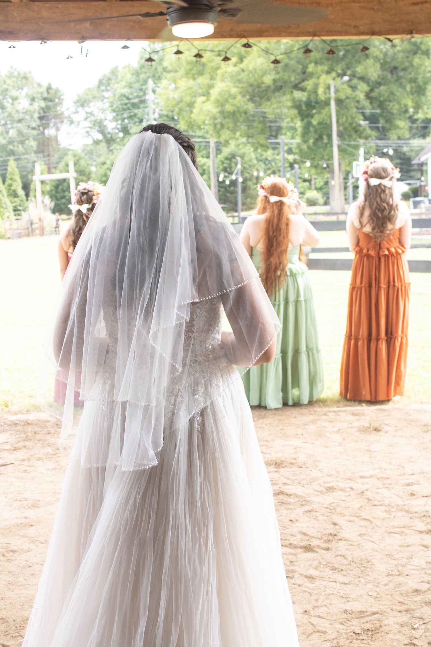 Bride in white veil standing with bridesmaids in colorful gowns at outdoor wedding venue.