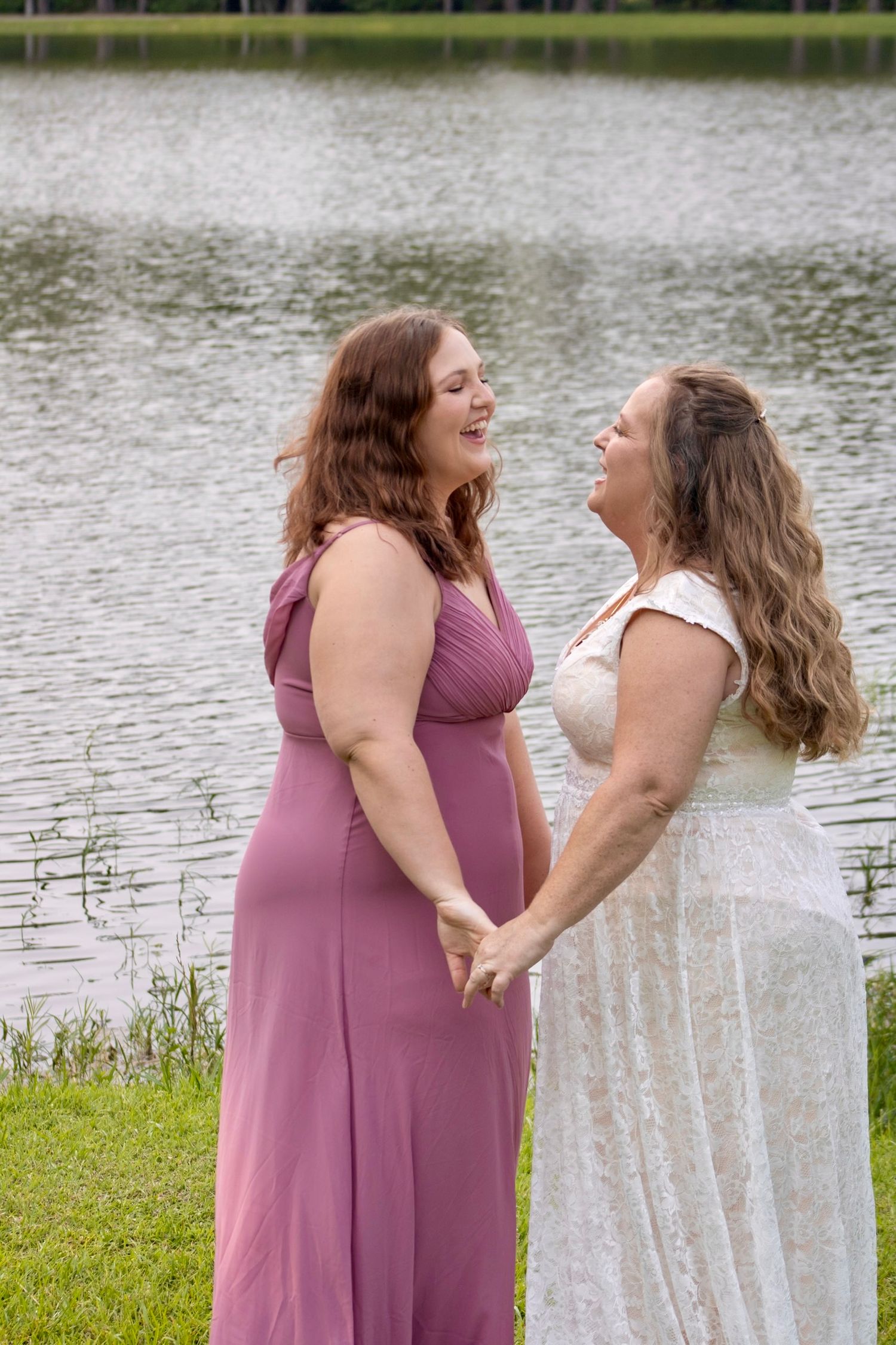 Two women in formal dresses hold hands and laugh together by a serene lake, one in white lace and one in mauve.