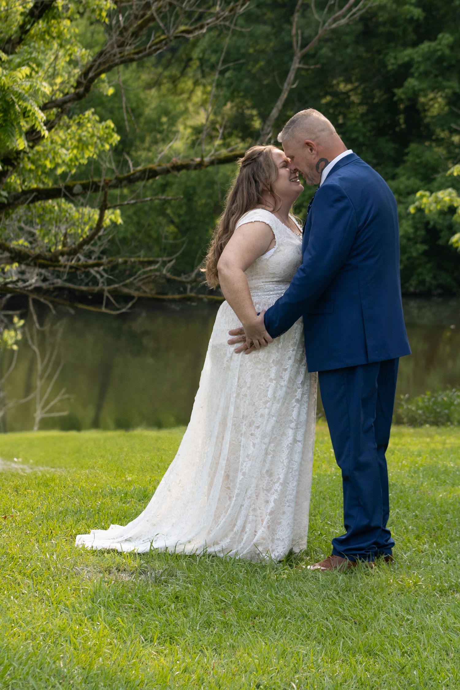Bride and groom share a romantic moment outdoors, with lush green trees and a pond in the background.