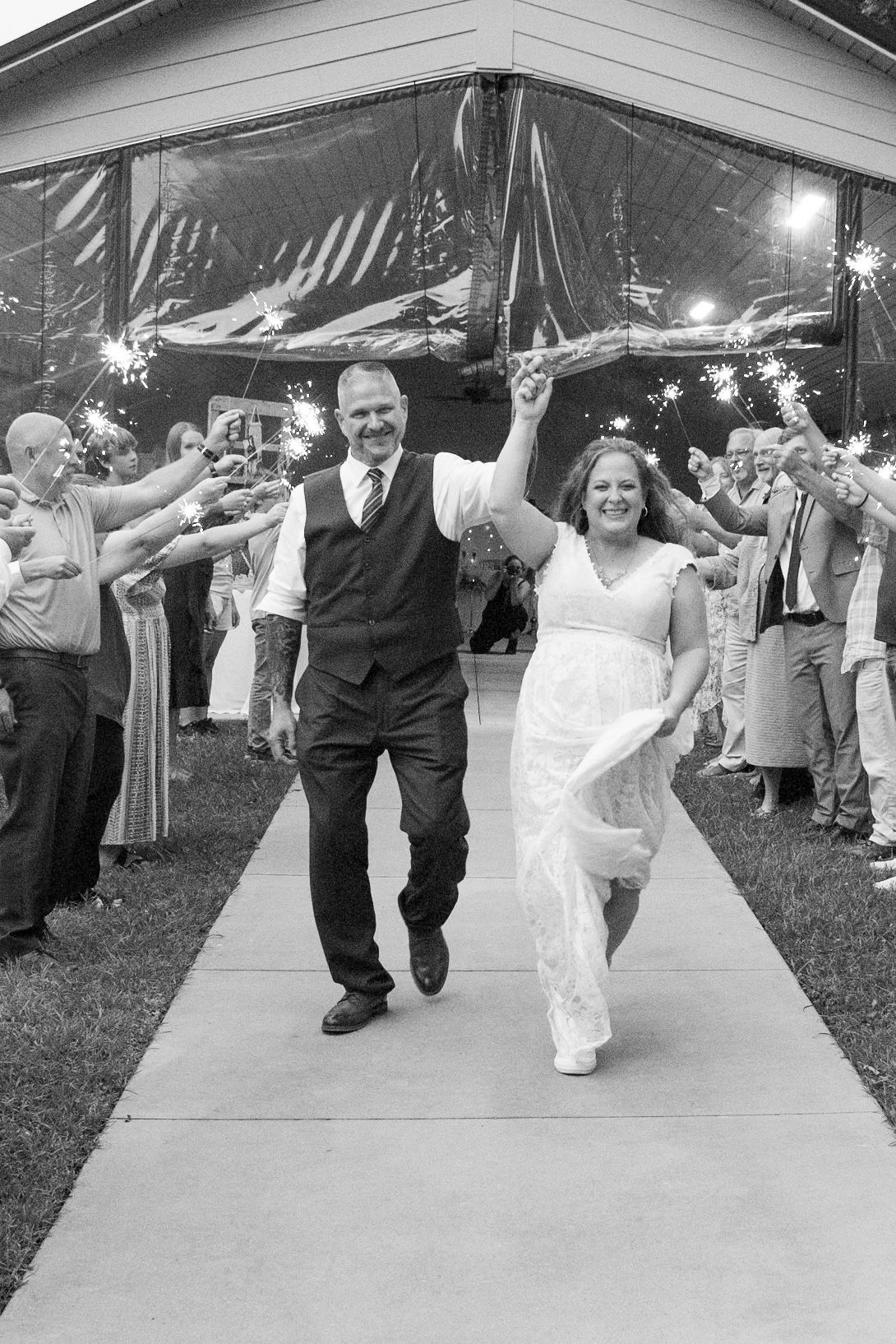 Black and white photo of a joyful bride and groom walking through a sparkler send-off at their outdoor wedding reception.