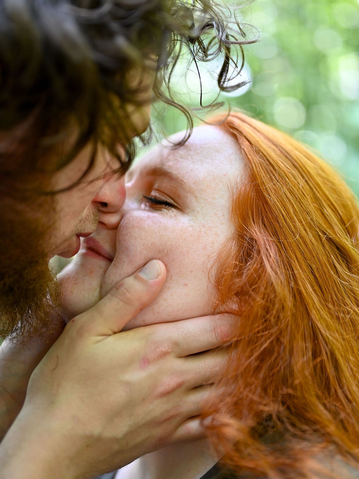 A man with a beard tenderly kisses a redhead woman with freckles outdoors in a lush green setting.