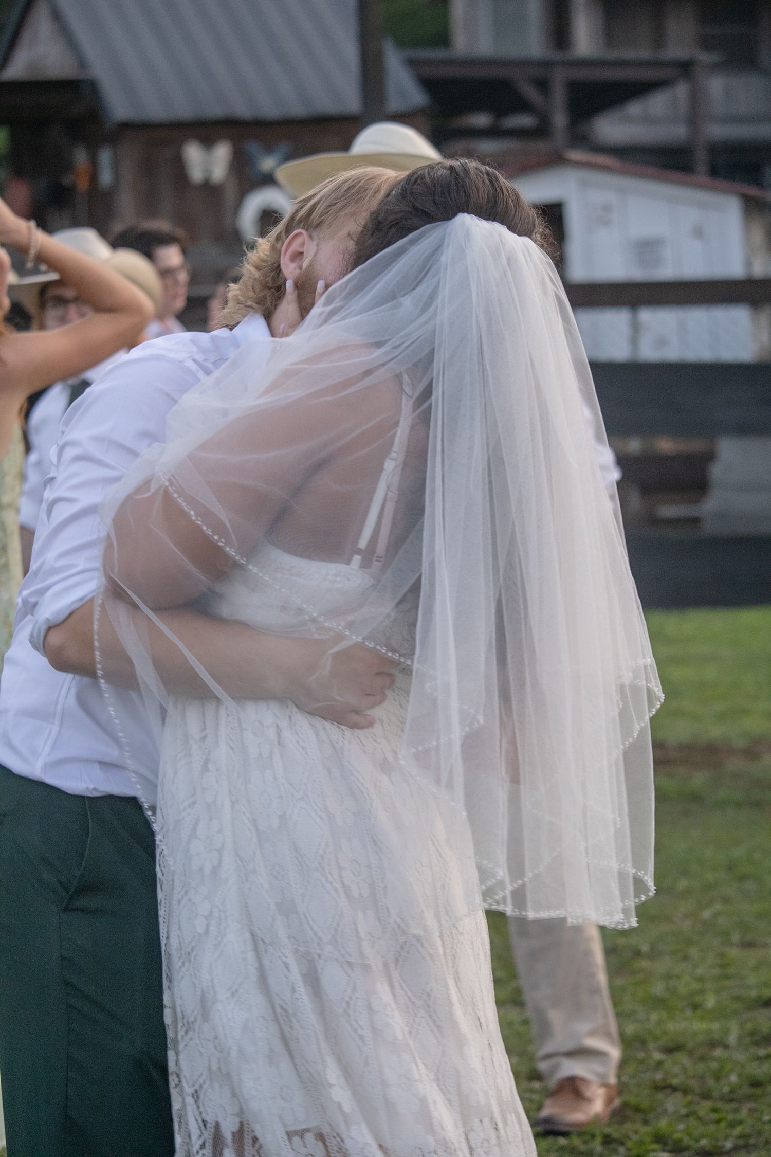 Bride in white lace gown and veil sharing emotional embrace with guest at outdoor rustic wedding venue.