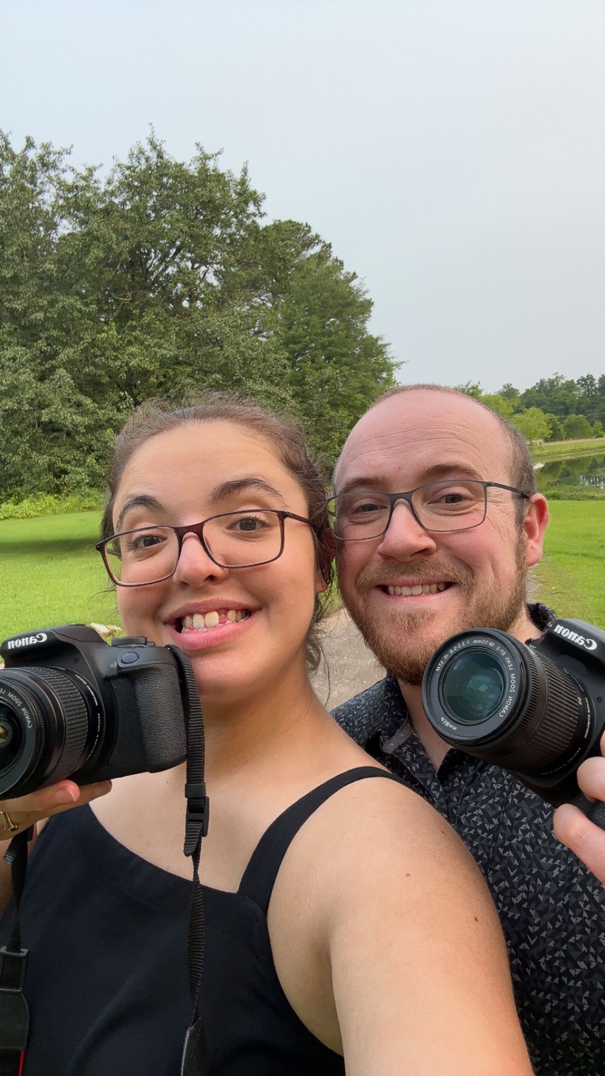 Two wedding photographers smiling and holding Canon DSLR cameras outdoors at a green park venue.