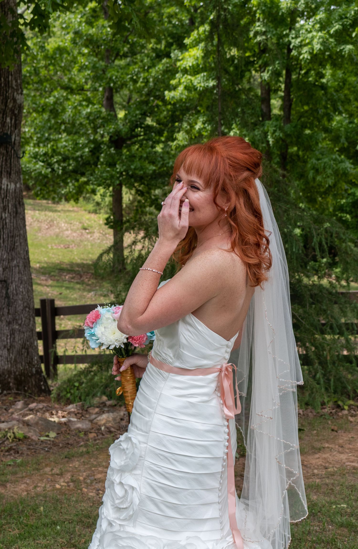 Emotional redhead bride in backless white gown with veil holds bouquet outdoors among trees.