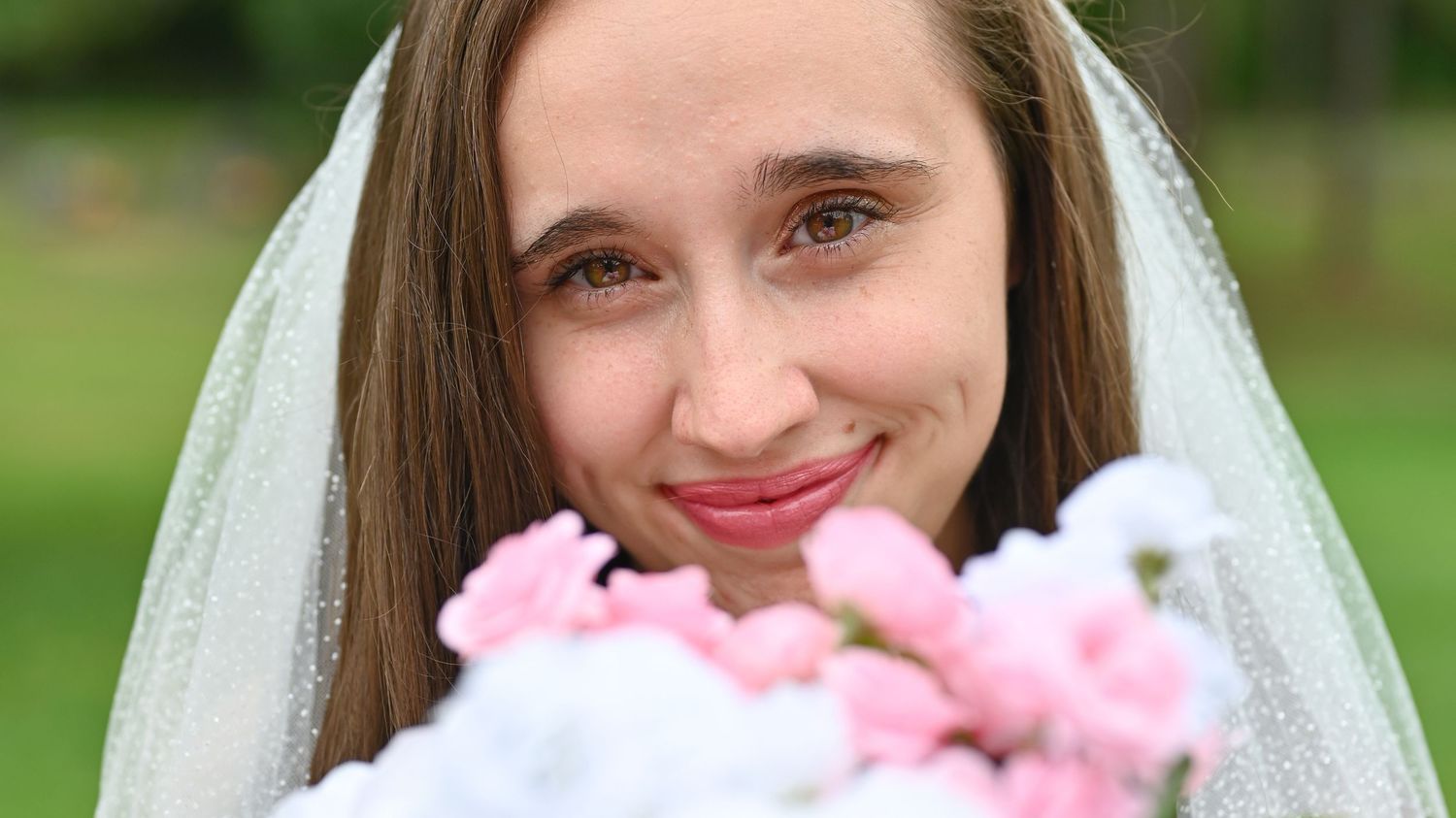 Smiling bride in white veil holding pink and white flower bouquet outdoors on her wedding day.
