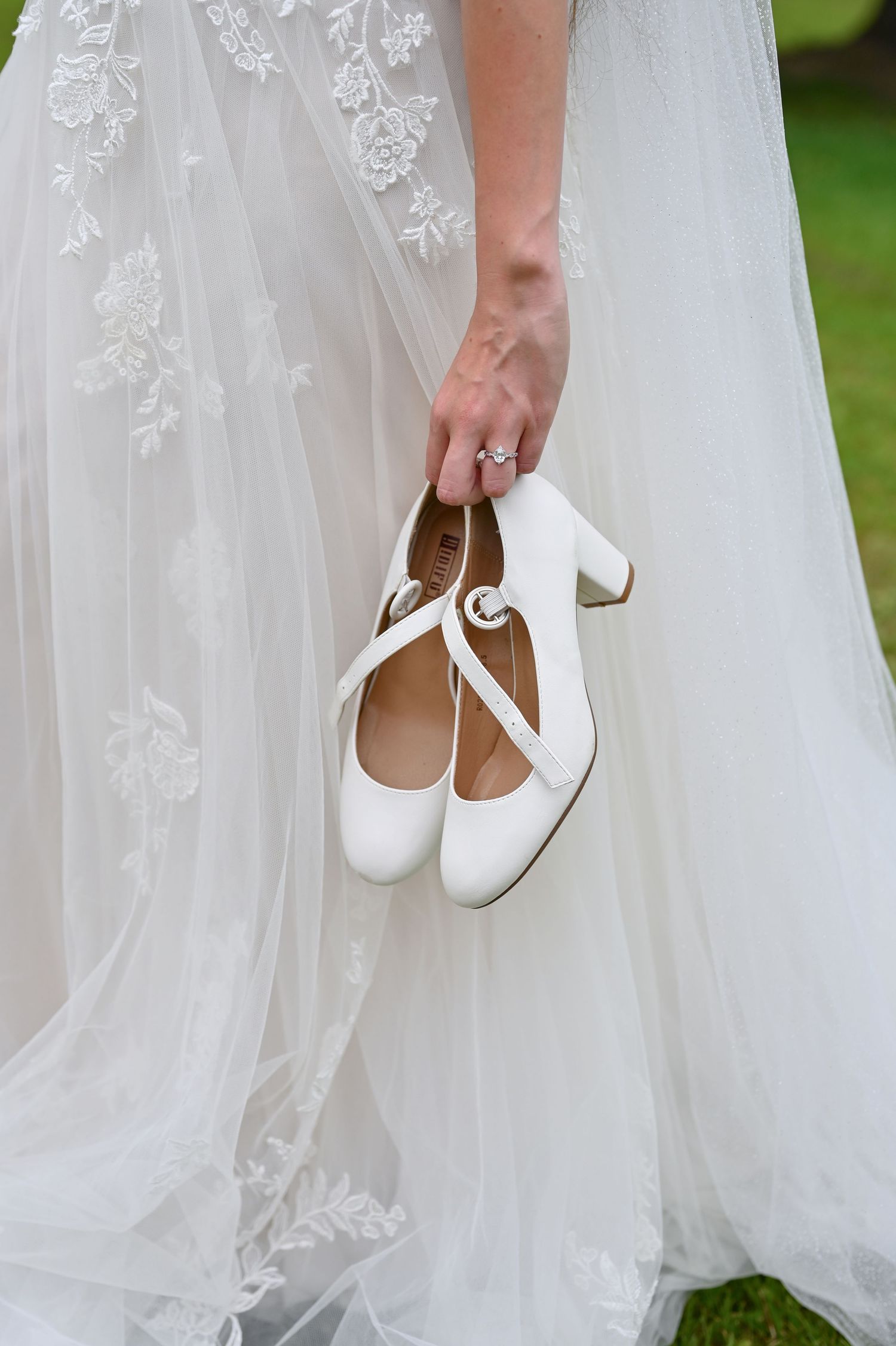 Bride in lace wedding gown holding white Mary Jane block heel bridal shoes with diamond engagement ring on her finger.