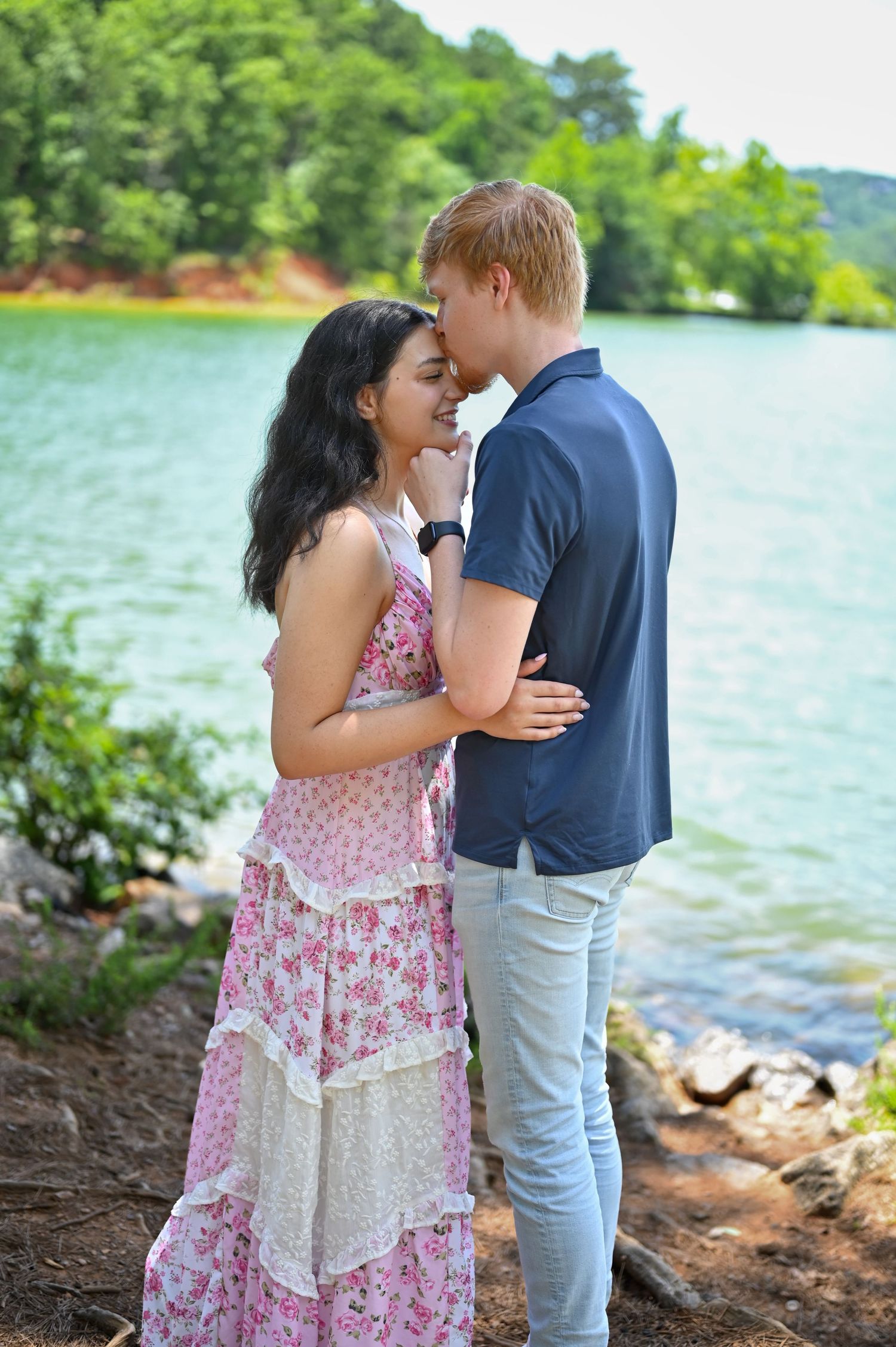 Couple sharing a romantic moment by a lakeside, woman in floral pink dress and man in navy shirt embracing.