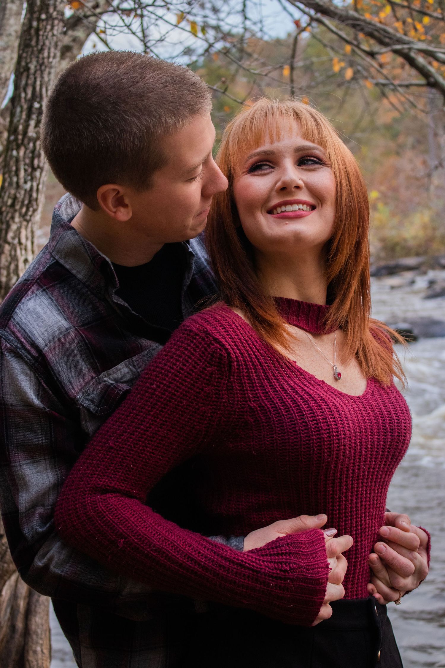A couple embraces outdoors in autumn, woman in burgundy sweater smiling warmly as man holds her near a forest stream.