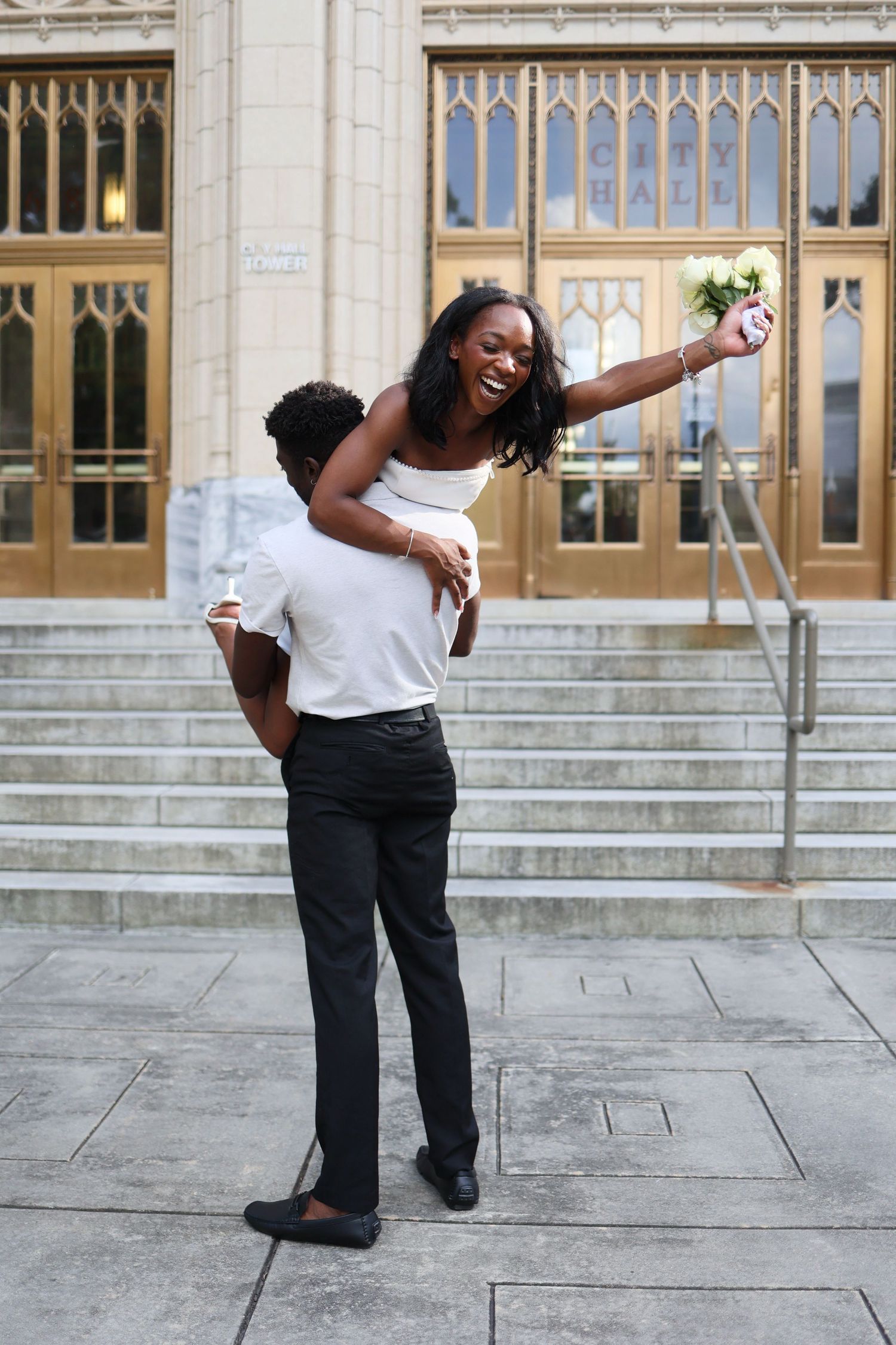 Joyful Black groom carrying bride piggyback outside City Hall, bride laughing and raising bouquet.