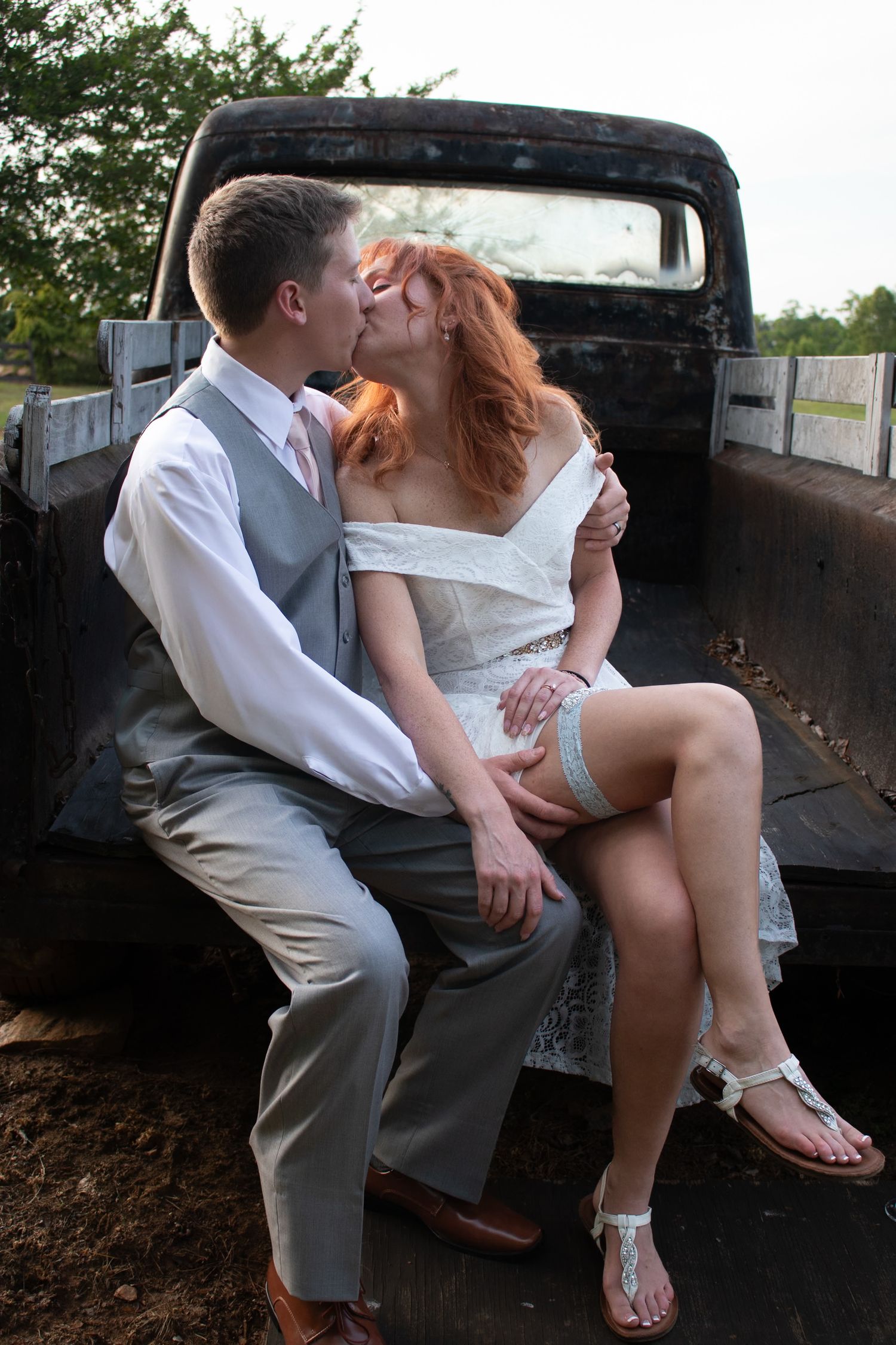 Newlywed couple sharing a romantic kiss while seated on the tailgate of a vintage rusty pickup truck.
