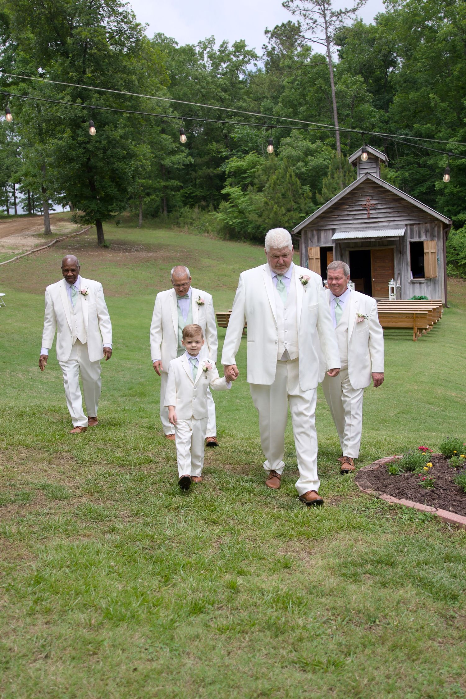 Groomsmen in white suits walk across green lawn with rustic chapel backdrop at outdoor wedding ceremony.