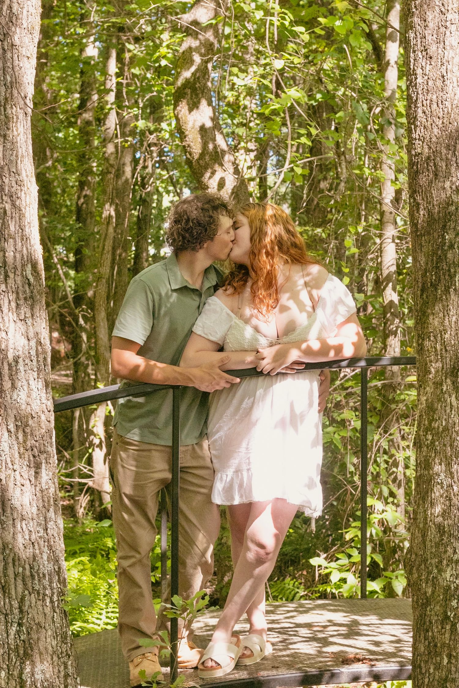 Couple kissing on a wooden bridge surrounded by lush green trees in a sunlit forest setting.