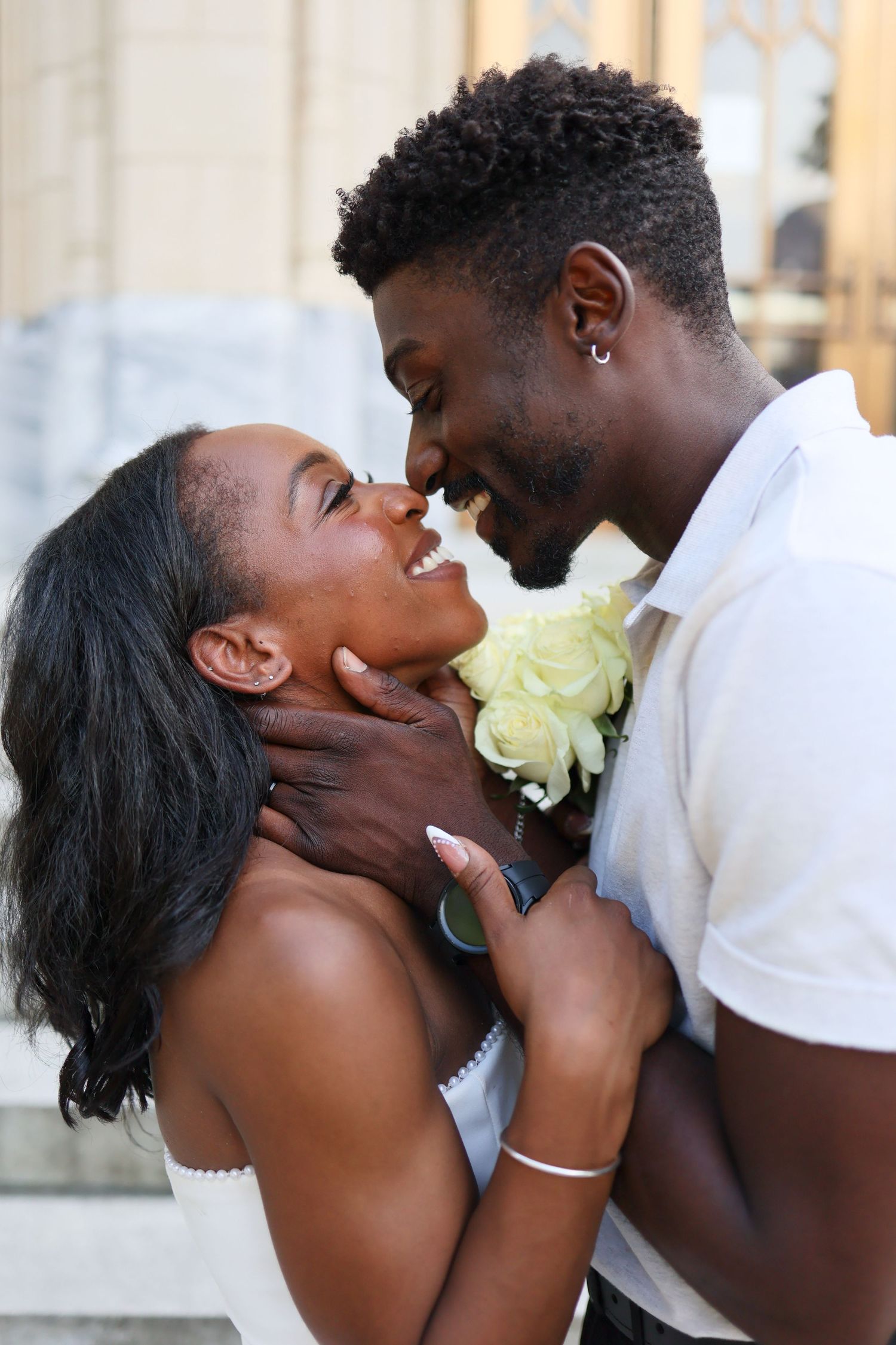 Happy Black couple almost kissing, groom tenderly holding bride's face with white roses between them.