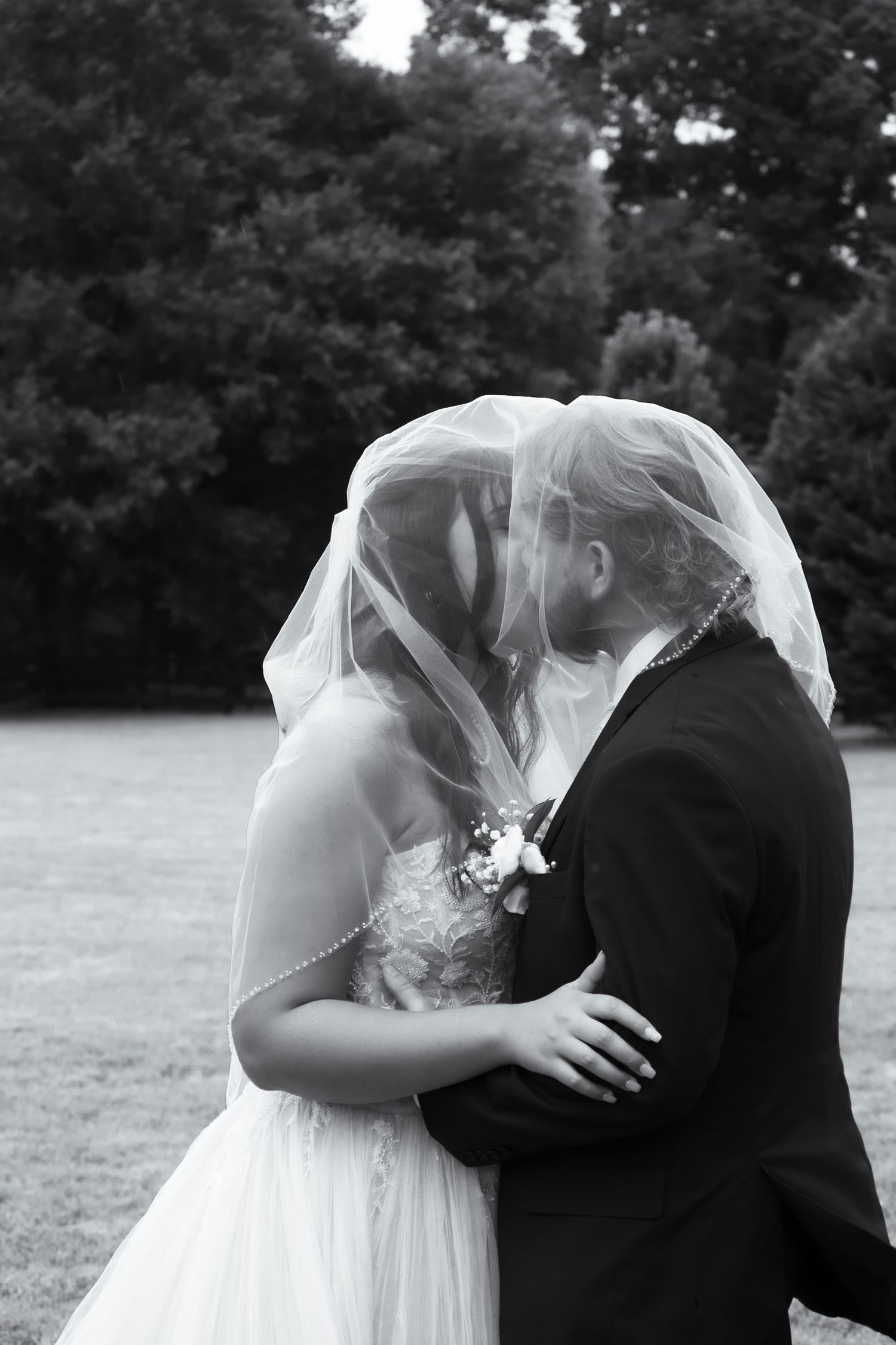 Black and white photo of bride and groom kissing under wedding veil outdoors.