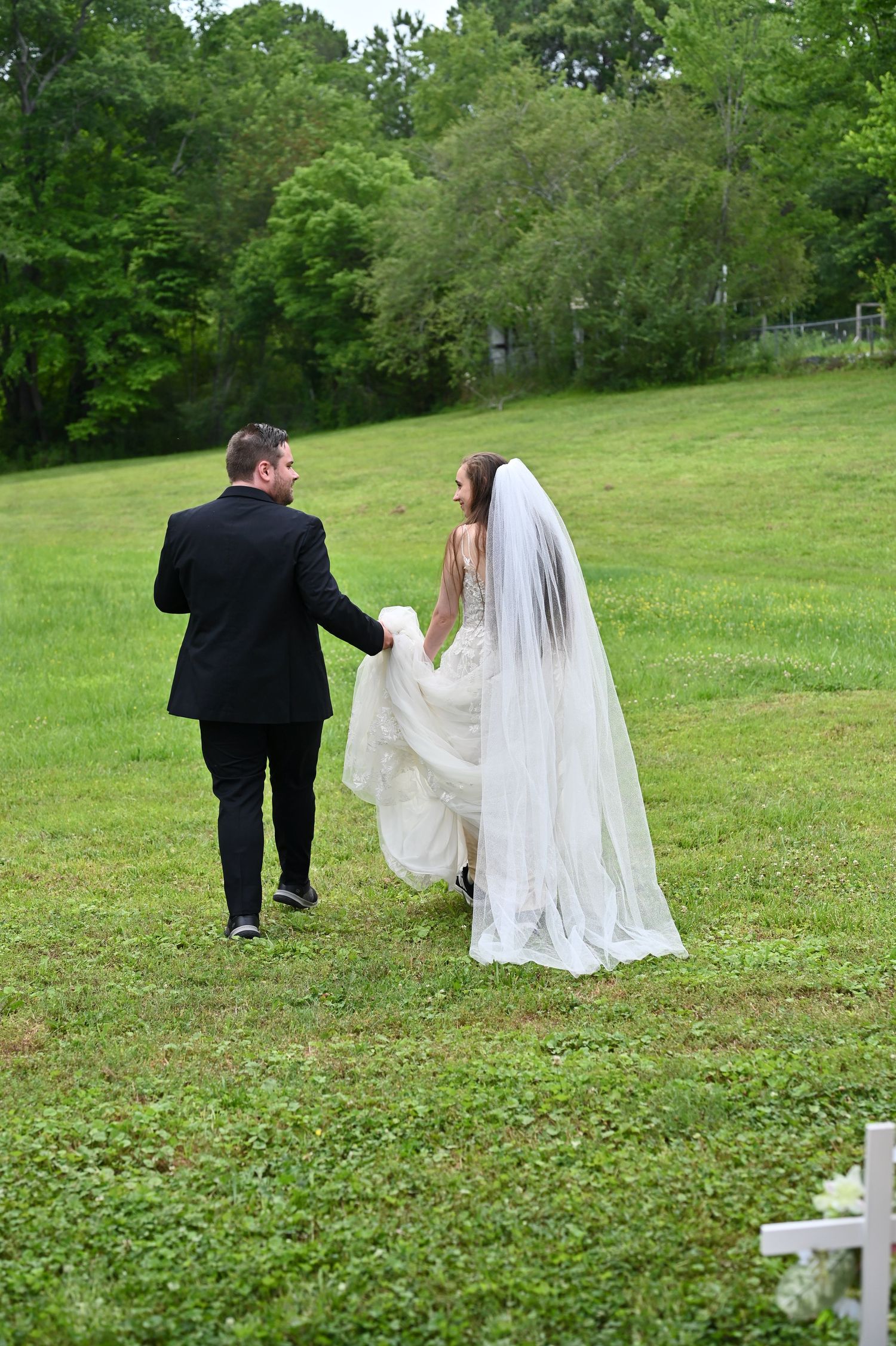 Bride in white gown and veil walks hand-in-hand with groom in black suit across lush green lawn.