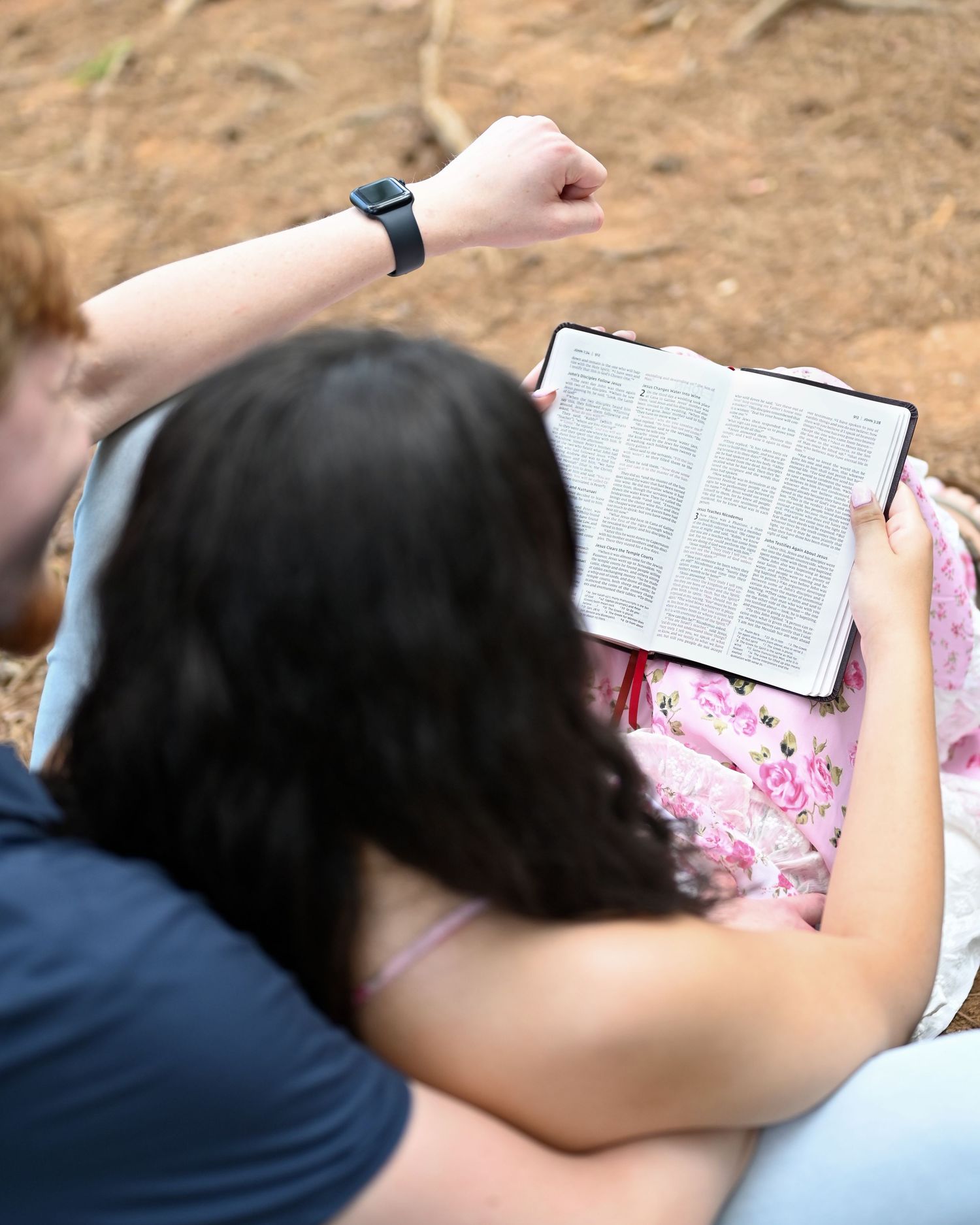 A couple sits outdoors reading a Bible together, the woman holding the open book while the man raises his fist.