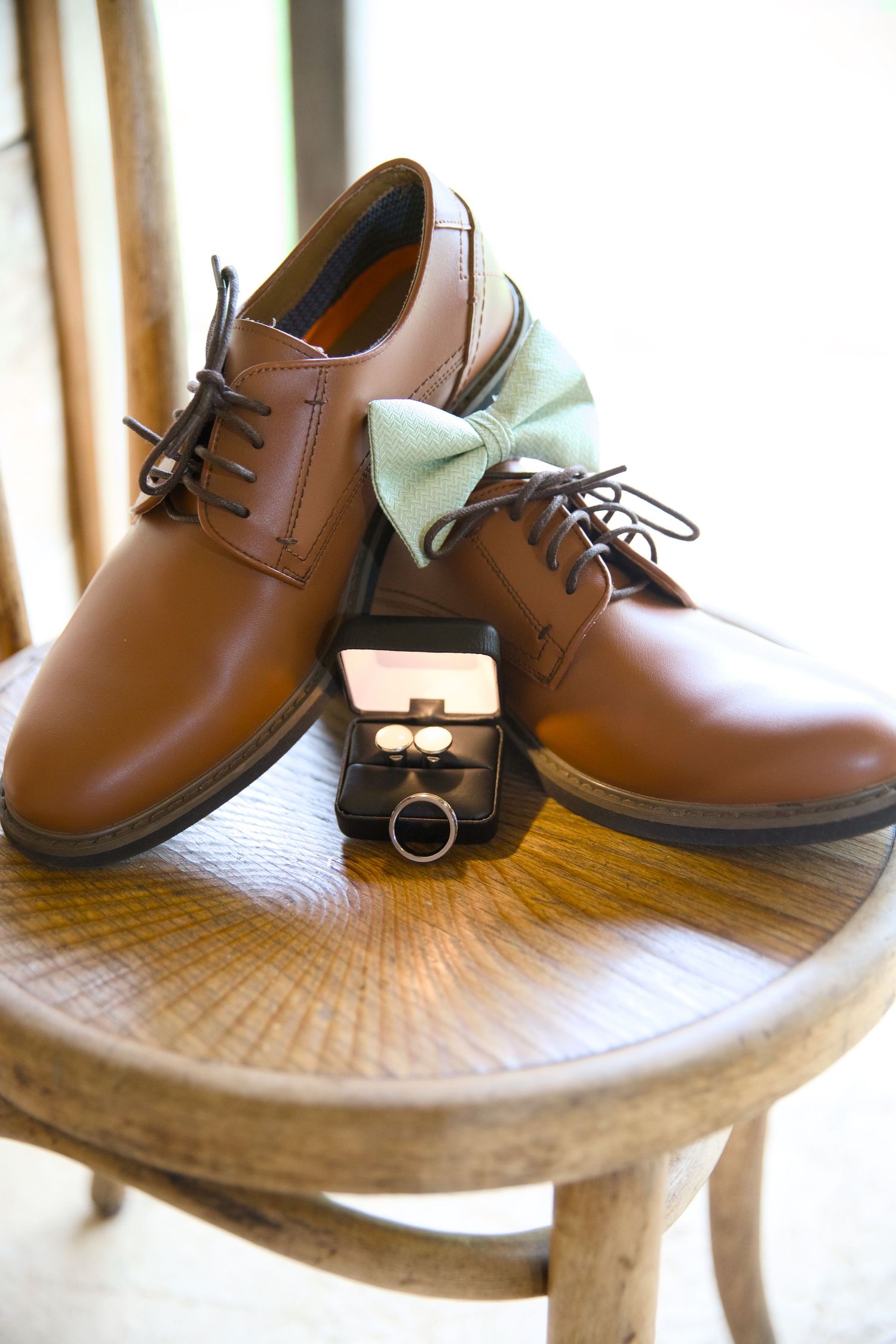 Brown leather dress shoes with mint bow tie and cufflinks on wooden chair for a groom's wedding day accessories.