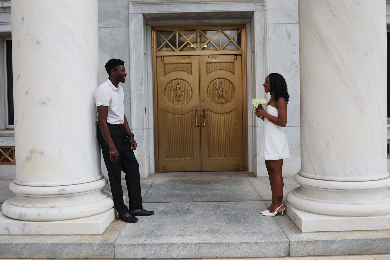 Couple in formal attire standing before ornate wooden doors flanked by marble columns.