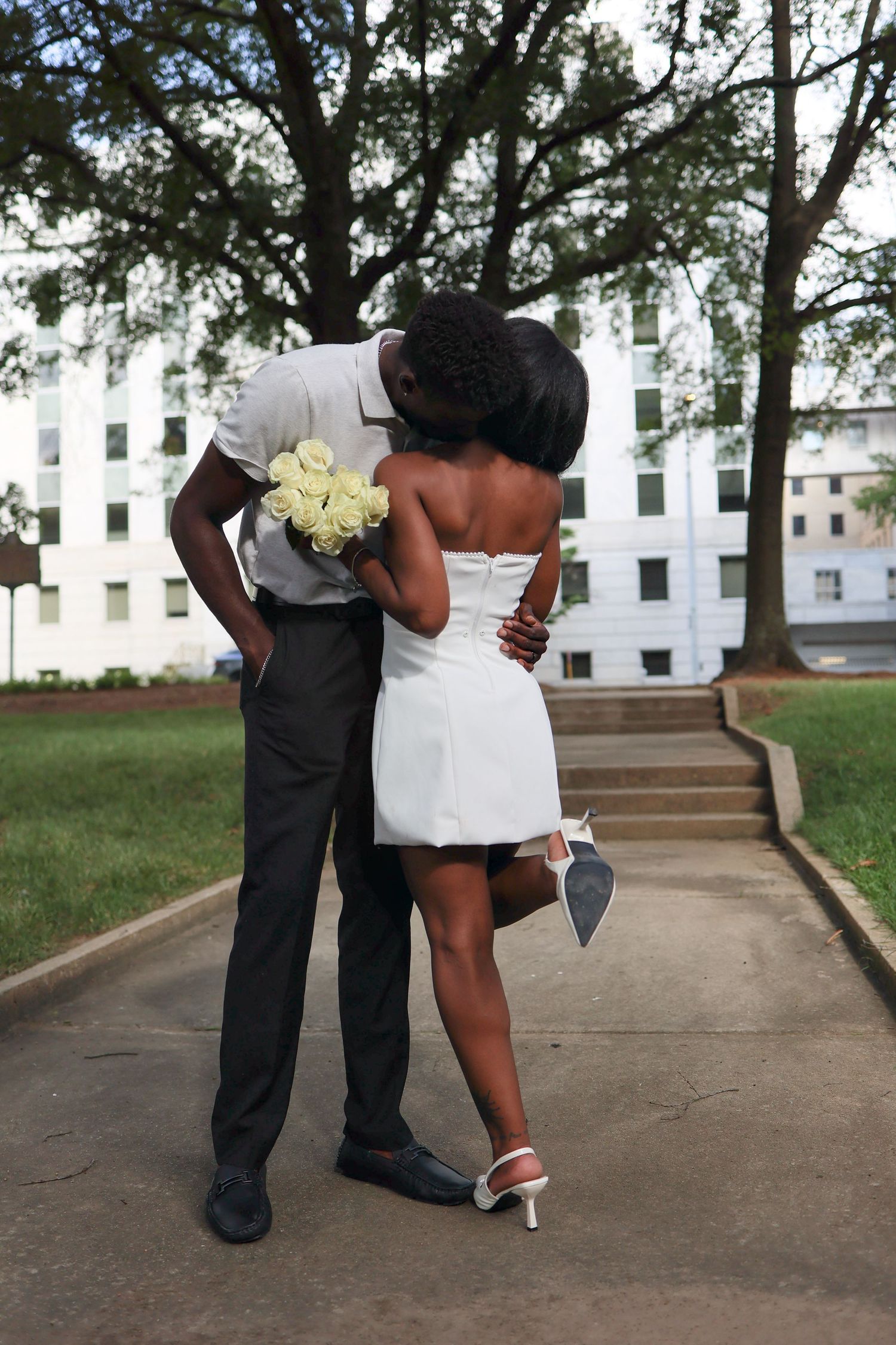 A couple shares a romantic kiss outdoors, the woman in a white mini dress holding heels, the man holding white roses.