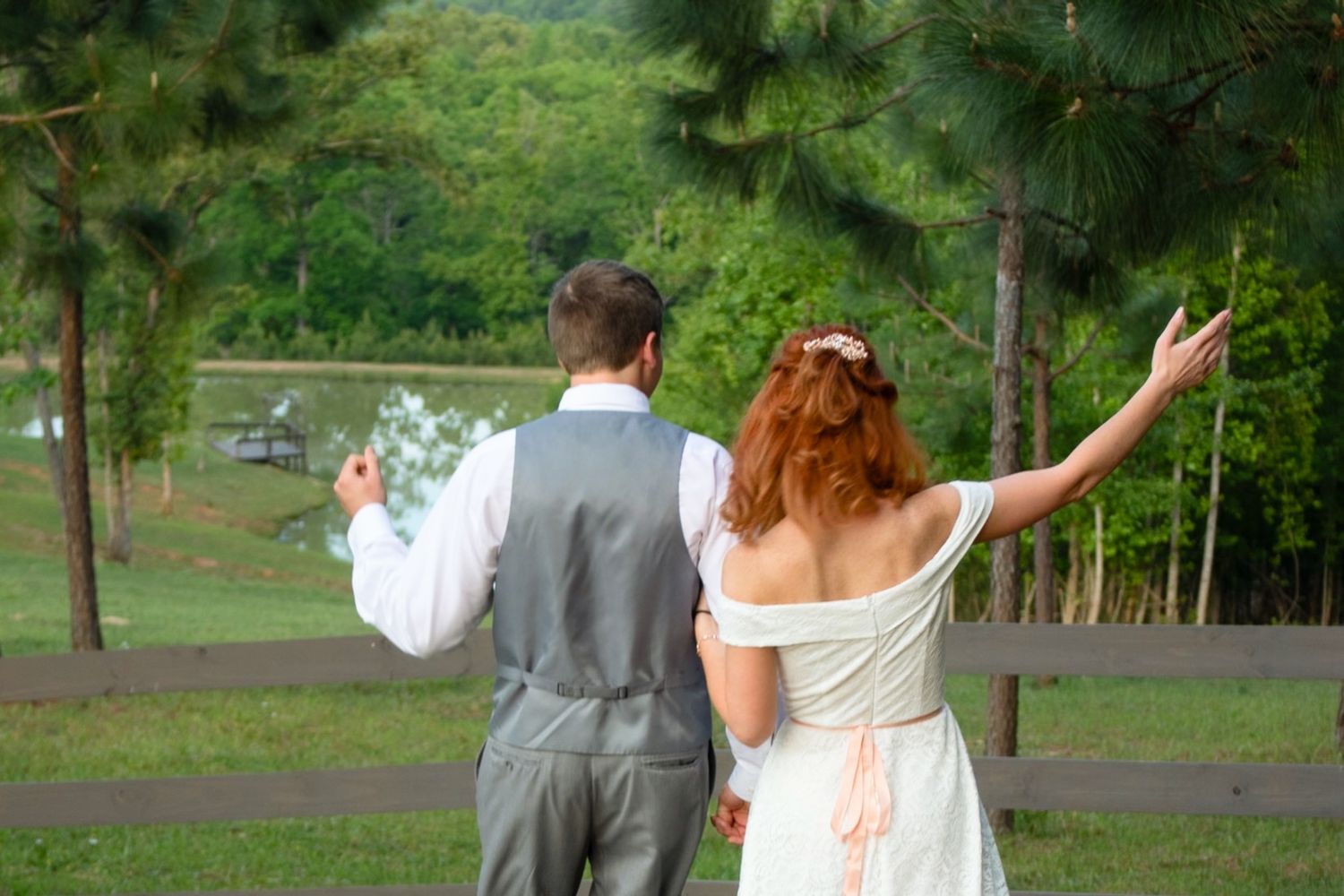 Bride and groom stand at a wooden fence admiring a lush green landscape on their wedding day.