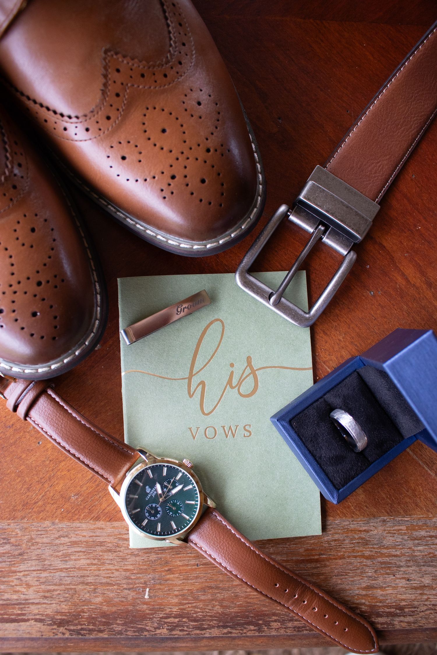 Groom wedding flat lay with brown leather dress shoes, belt, watch, ring, cufflinks, and his vows card on wood.
