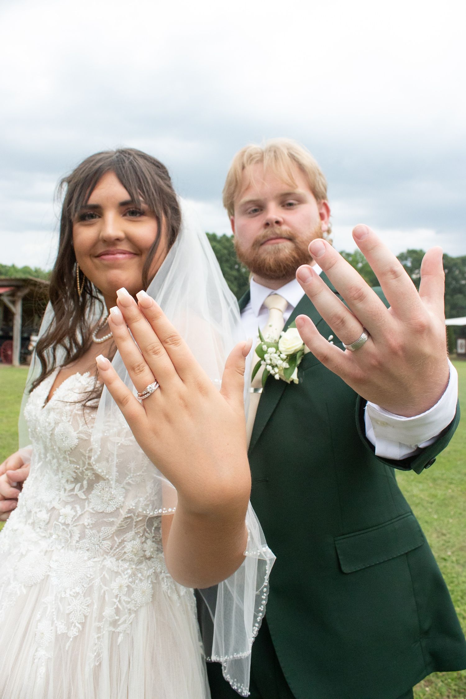 Newlywed couple showing off wedding rings on their hands, bride in white lace gown with veil, groom in green suit.