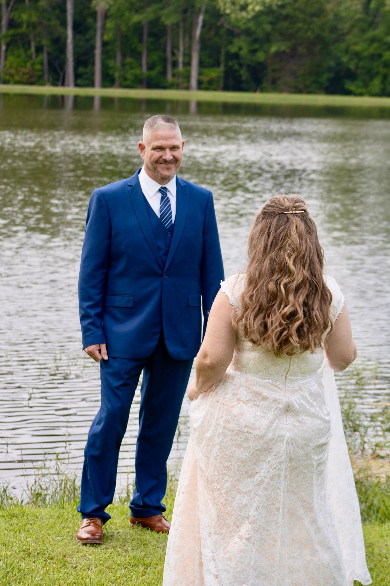 A bride in a white lace gown faces a smiling groom in a navy suit by a serene lake surrounded by trees.