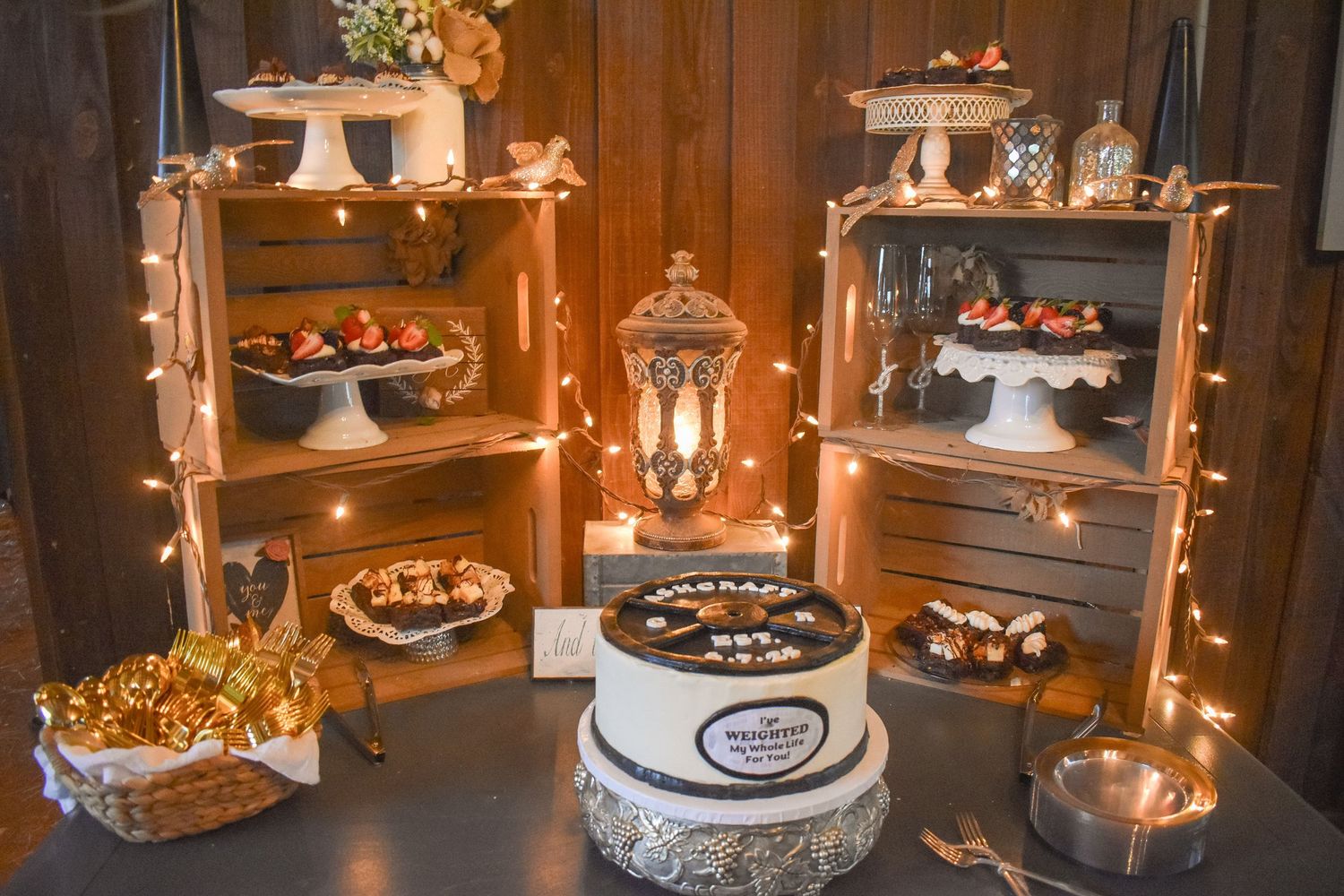 Rustic wedding dessert table with wooden crate shelves, fairy lights, and decorated cake.