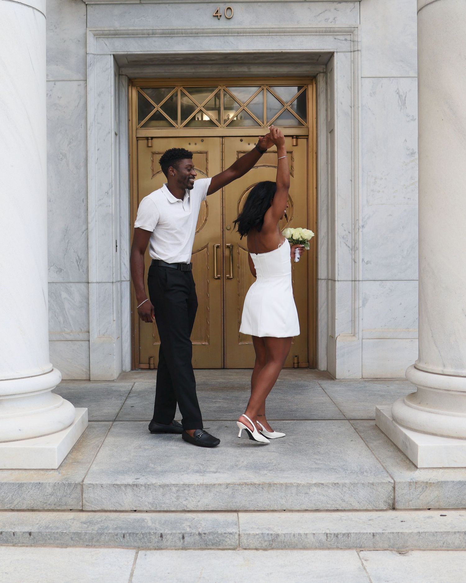A couple dances joyfully on marble steps before golden doors, the woman in a white dress holding a bouquet.