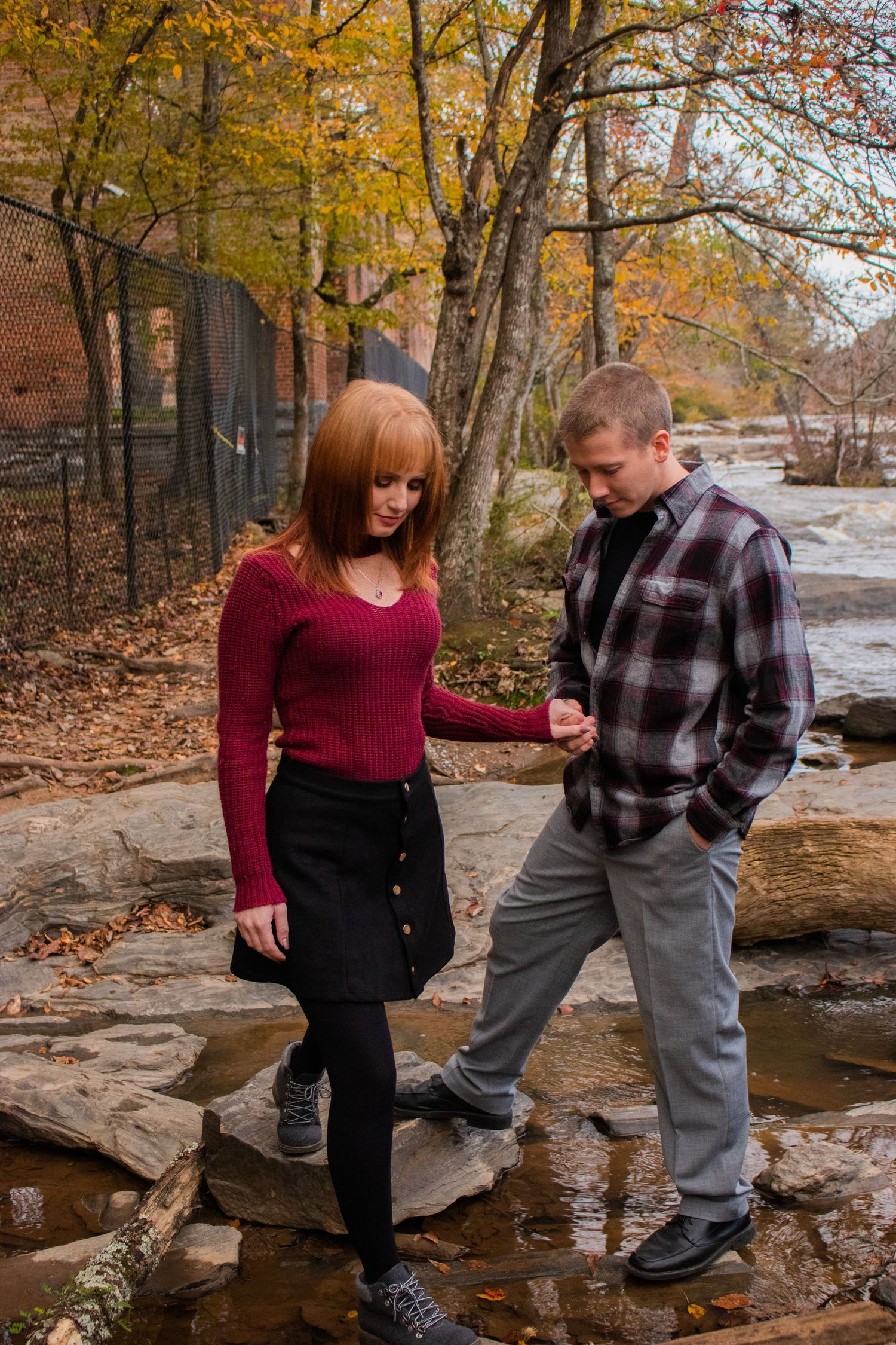 Couple holding hands on rocky path by autumn stream, woman in red sweater and black skirt, man in plaid shirt.