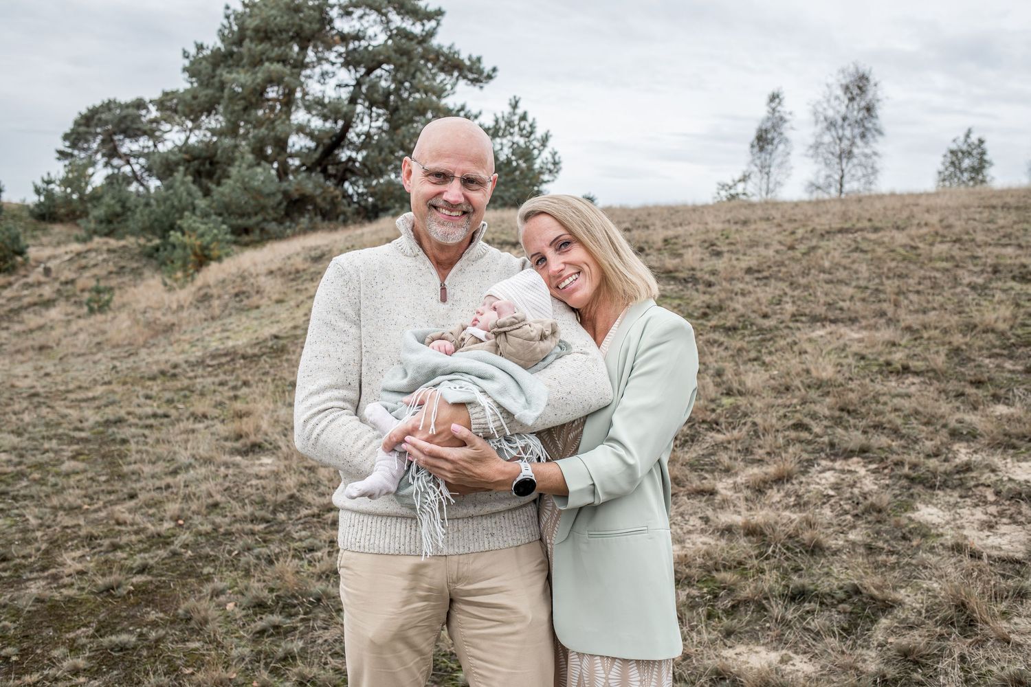 Smiling couple proudly holding newborn baby outdoors on grassy hillside with trees.