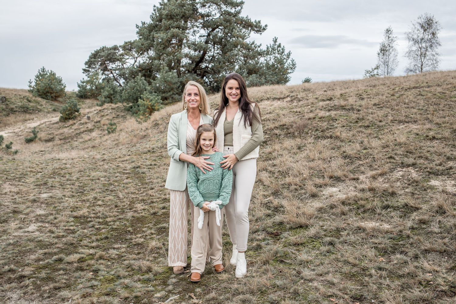 Three generations of women pose outdoors on a grassy hillside with pine trees in the background.