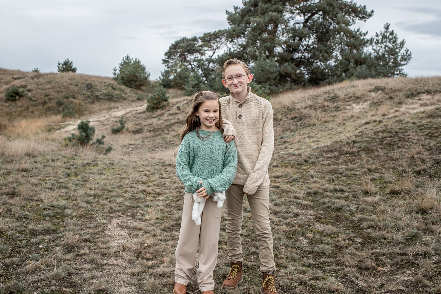 A young girl and teenage boy smile together outdoors on a grassy hill with pine trees.
