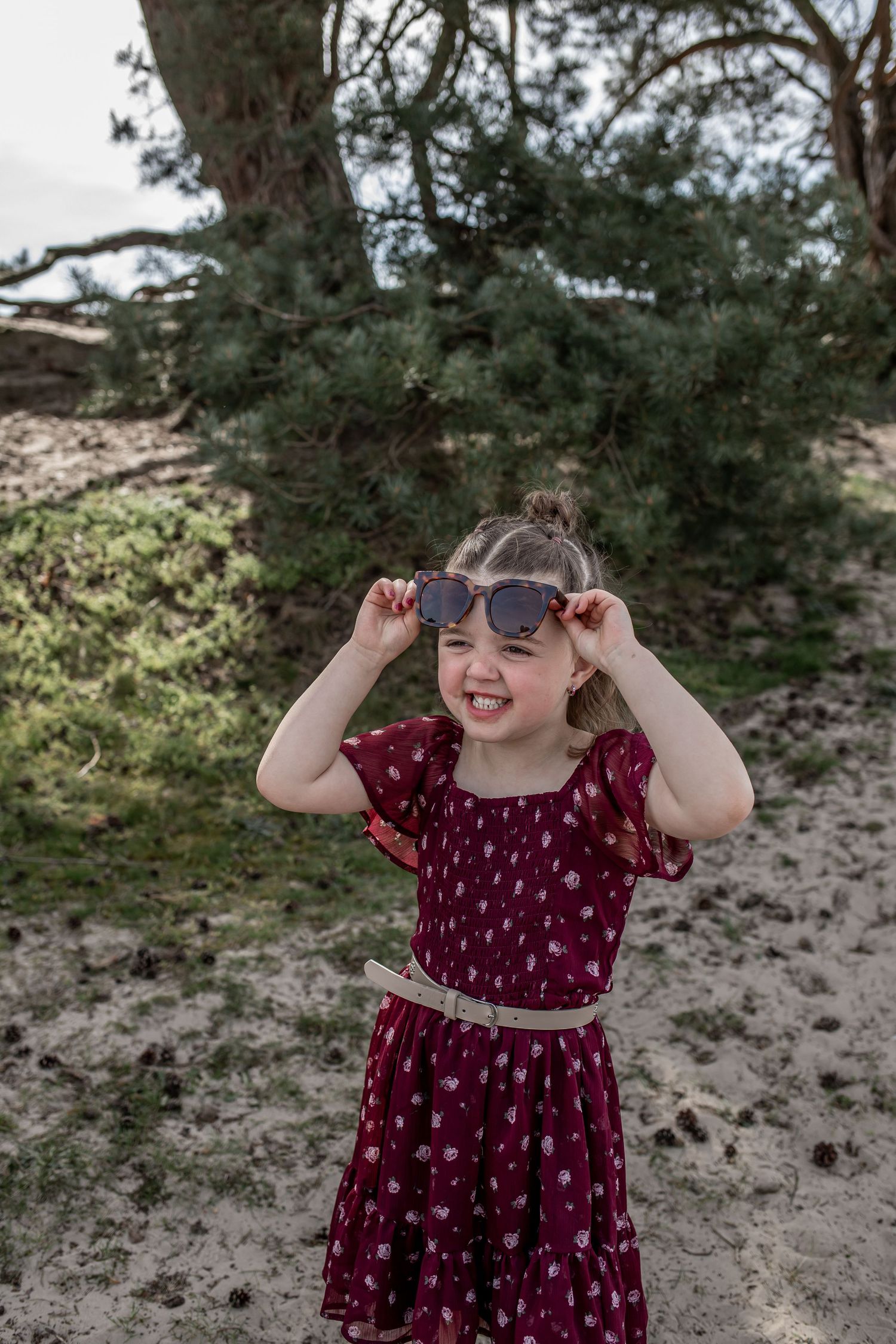 Young girl in red floral dress holding sunglasses, smiling outdoors near sandy ground and green pine trees.