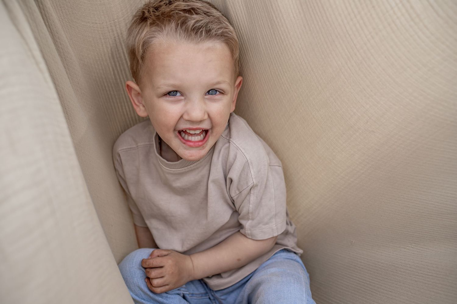 Happy young boy with blonde hair laughing while sitting between couch cushions wearing a beige shirt and jeans.