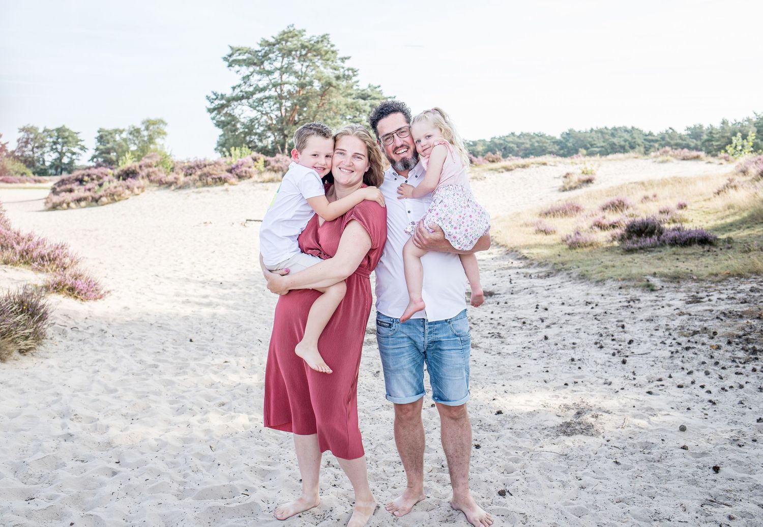 A joyful family of four poses barefoot on a sandy heath landscape with blooming heather in the background.