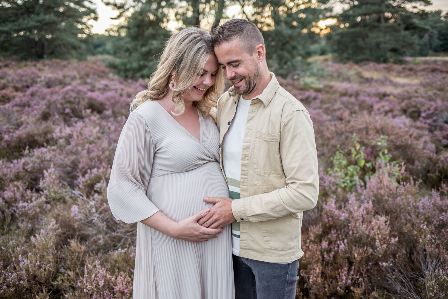 A couple smiles tenderly while the woman cradles her pregnant belly in a blooming heather field.