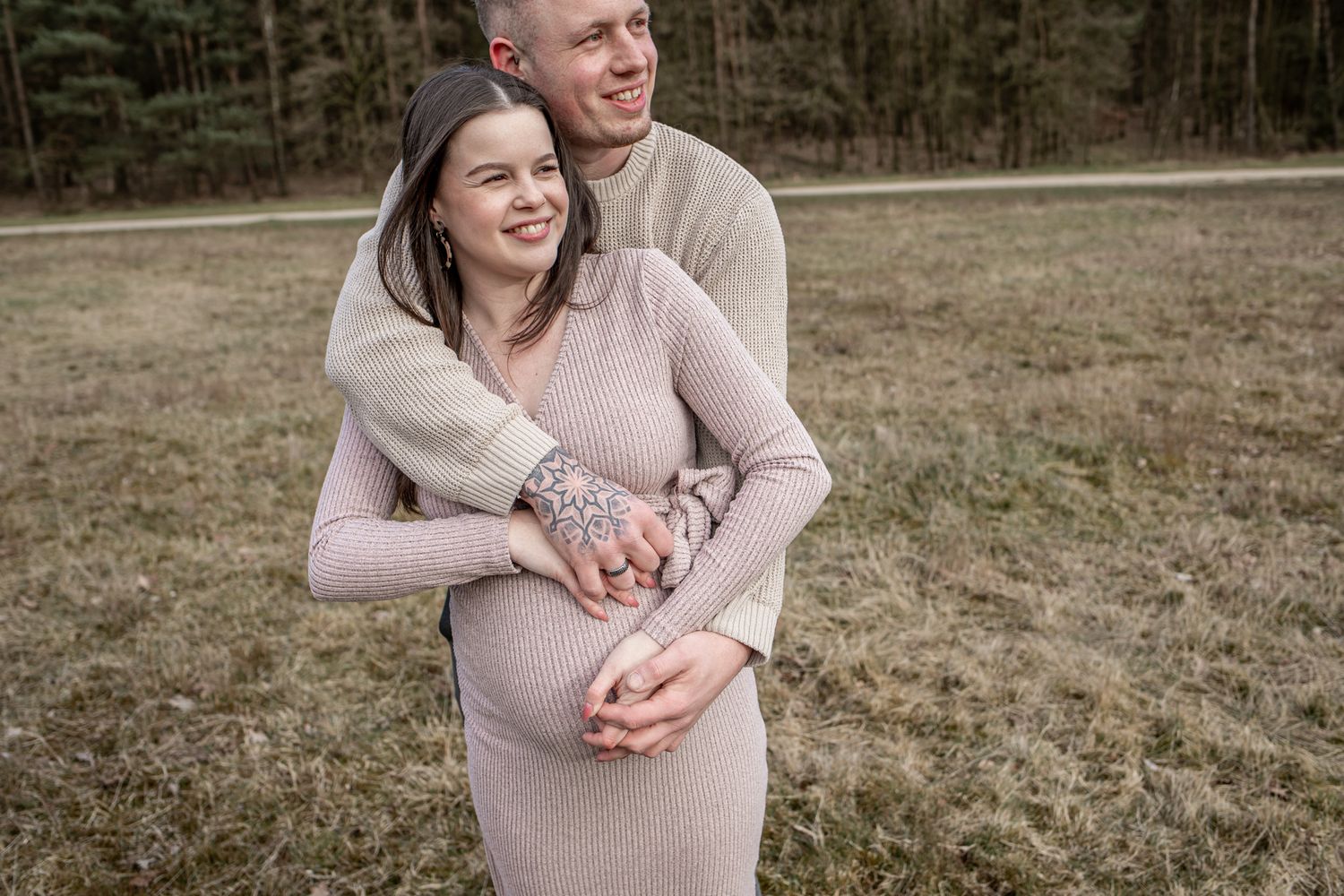Smiling pregnant woman in beige dress embraced by partner in grassy field.