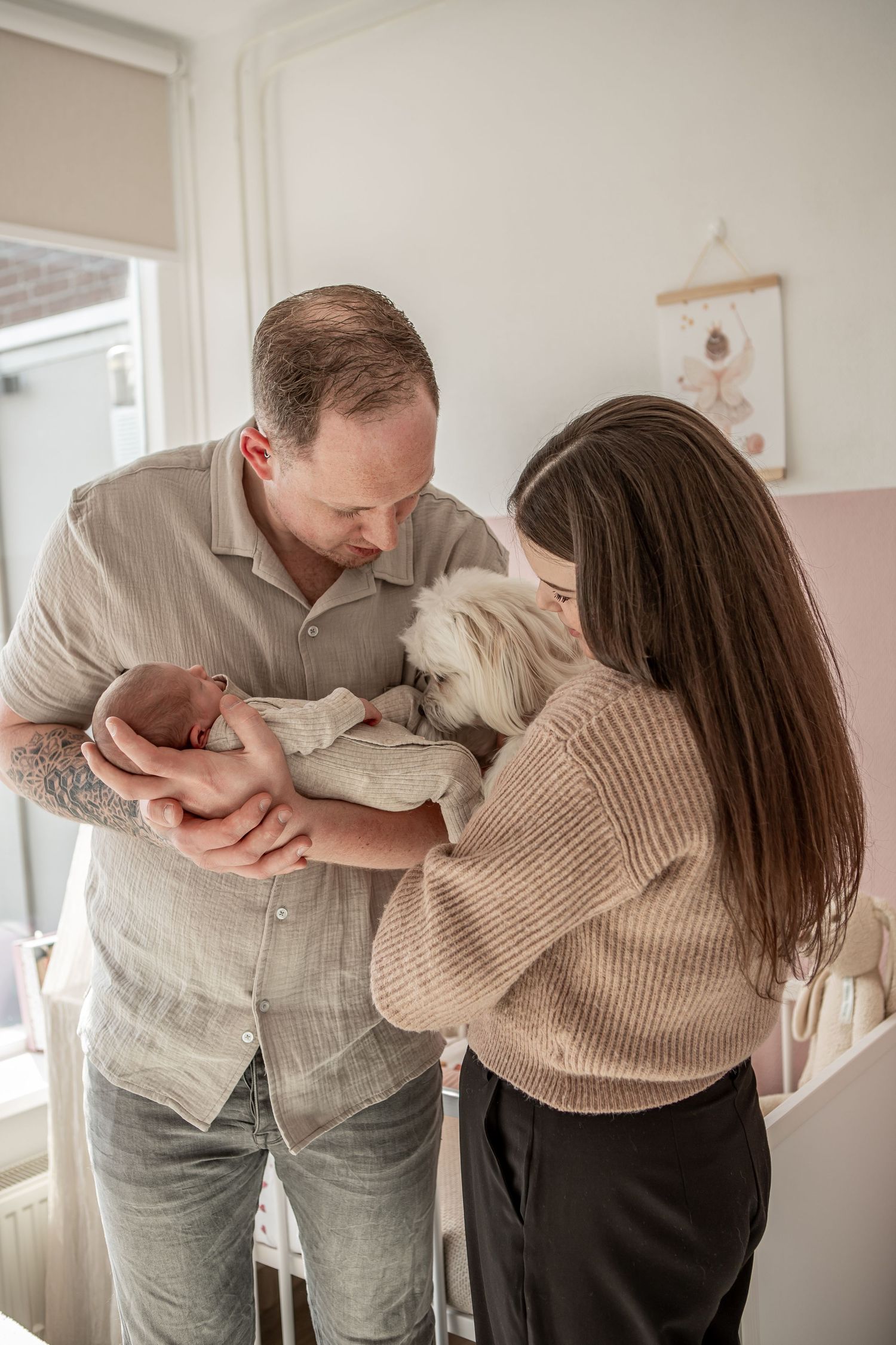 New parents and their small white dog admiring their swaddled newborn baby in a nursery.